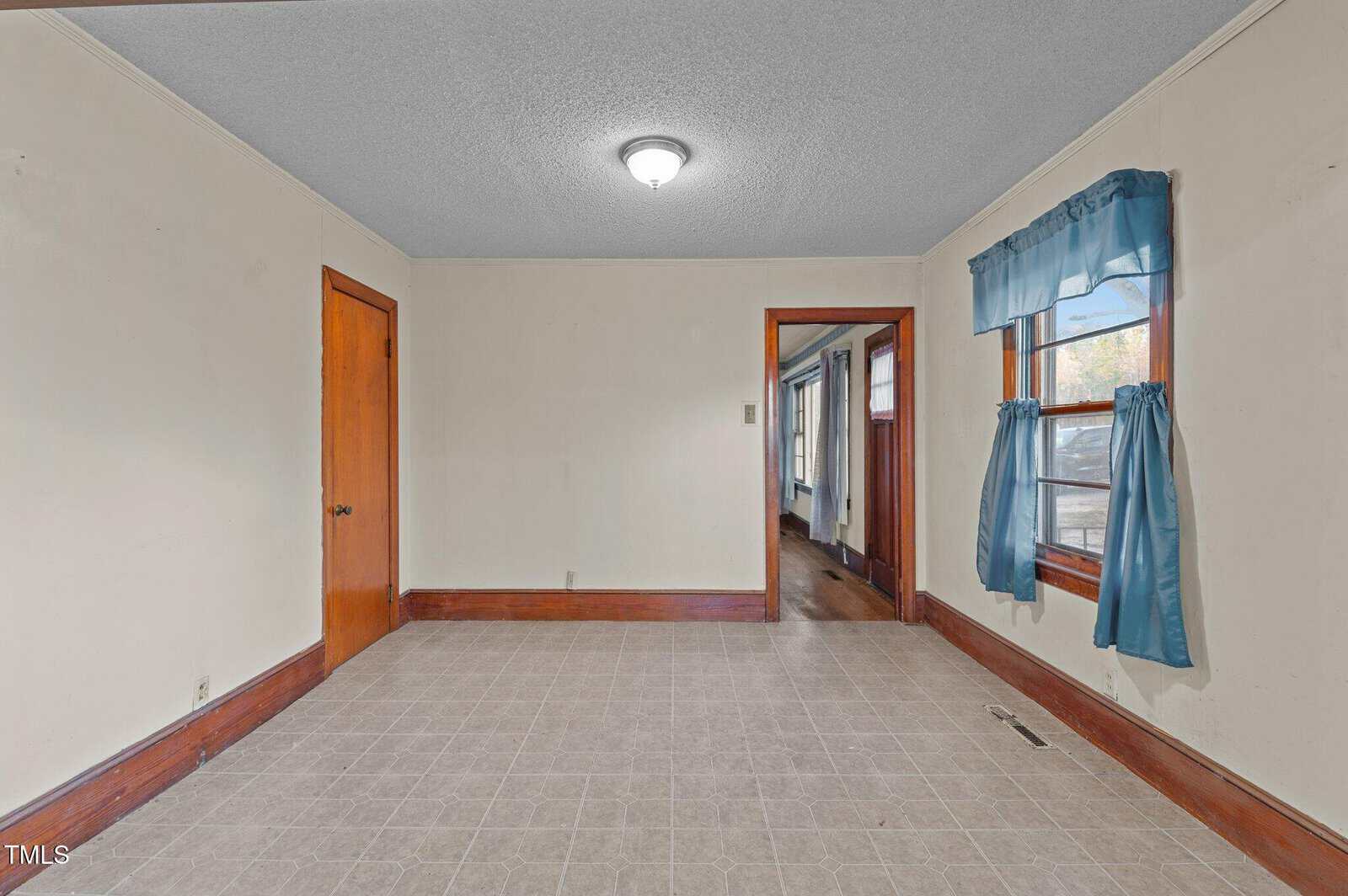An empty room with light-colored walls and a tiled floor. A door is on the left, and a window with blue curtains is on the right. A doorway leads to another room. The ceiling has a mounted light fixture.