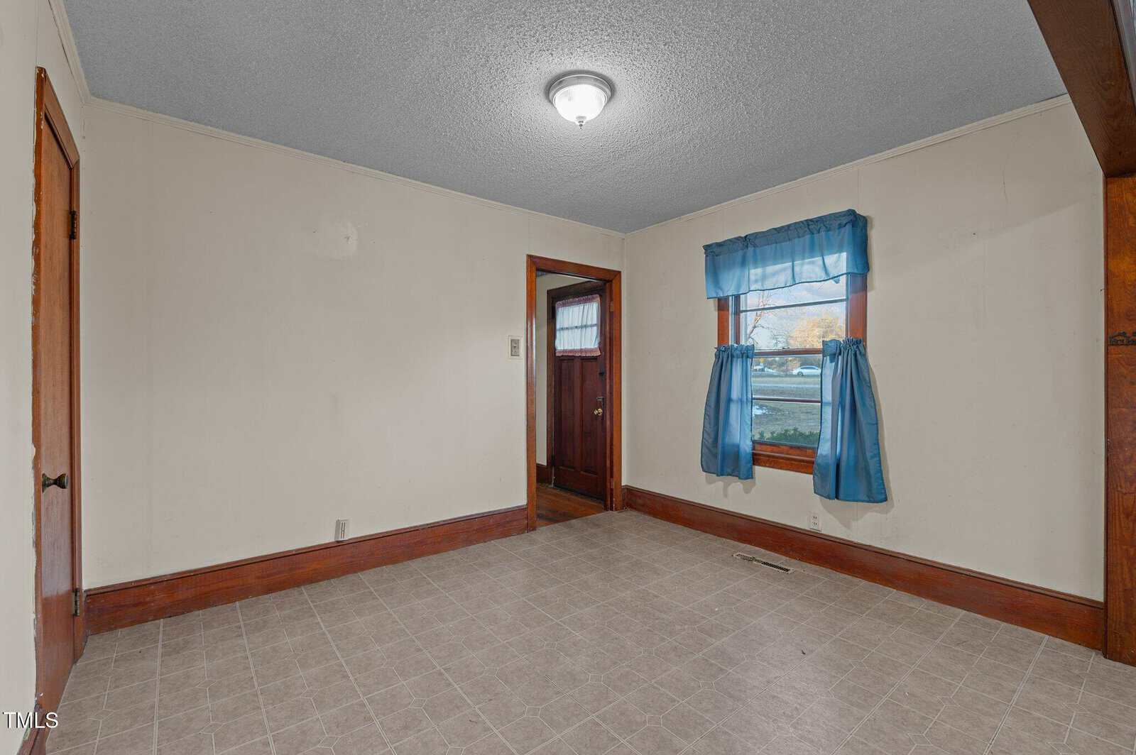 An empty room with beige walls and gray tile flooring. A window with blue curtains is on the right, and a wooden door to the outside is at the back. A light fixture is on the ceiling, and wooden trim borders the floor and doorway.