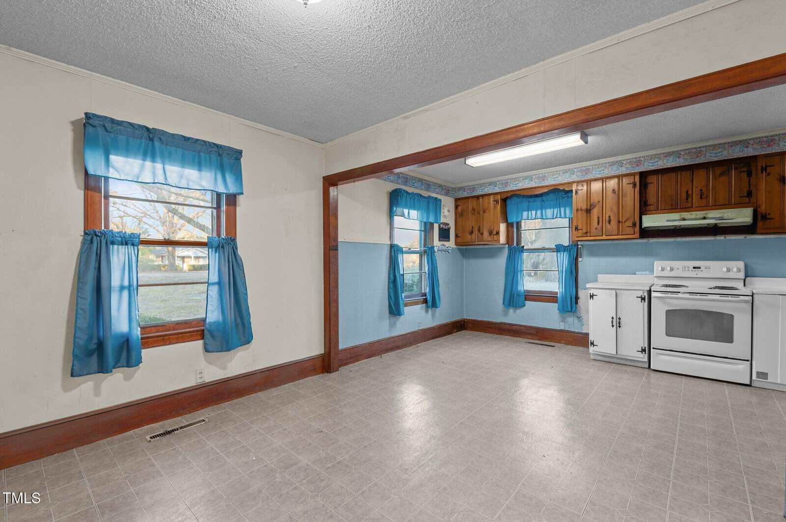 An empty room with a tiled floor features two windows with blue curtains. The room is partially divided by a wooden frame, leading to a small kitchen area with wooden cabinets. A white oven and range are positioned against a wall under a fluorescent light.