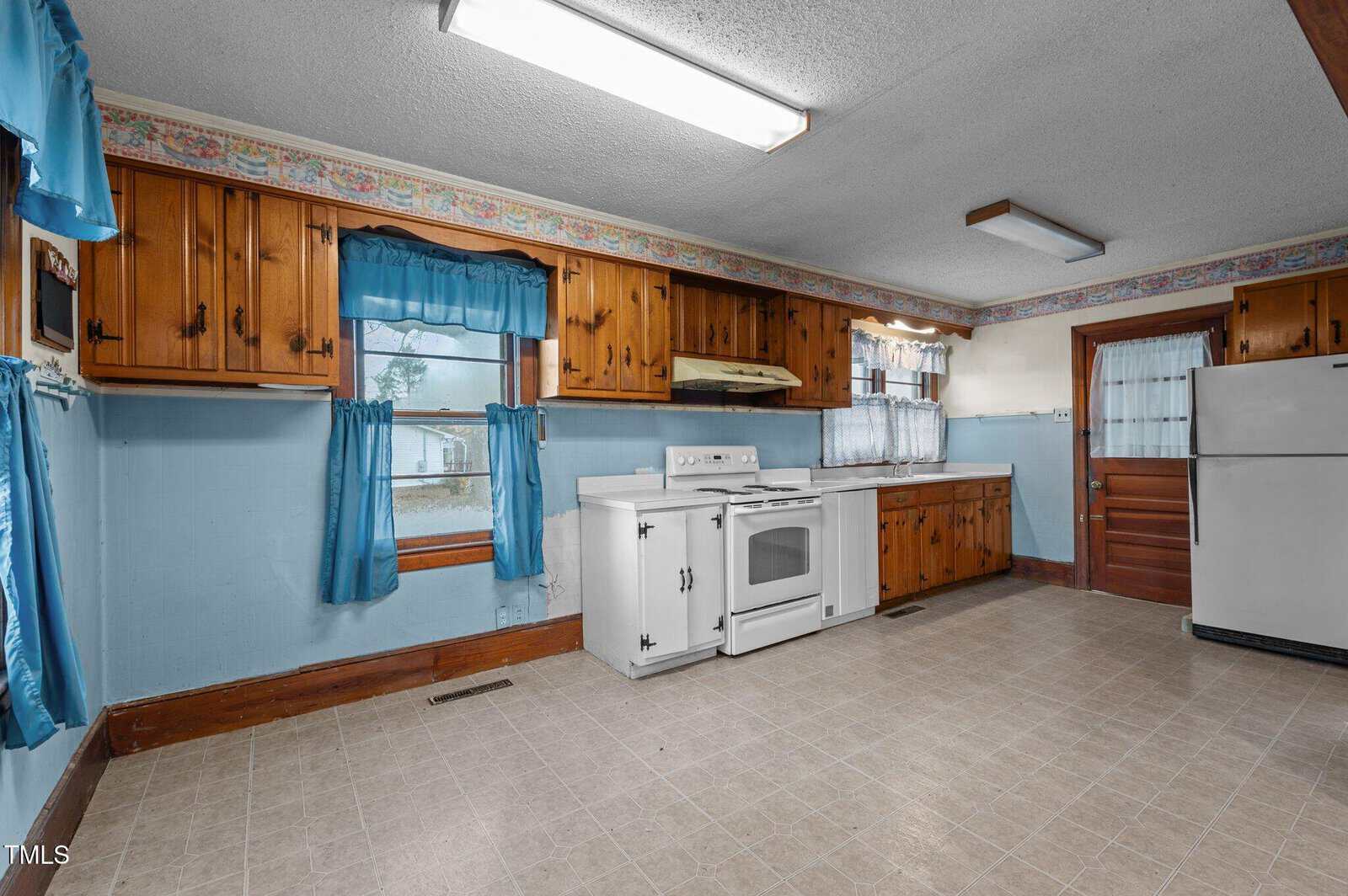 A vintage kitchen featuring wooden cabinets, a white stove, and refrigerator. The room has blue curtains and wallpaper border, with linoleum flooring. A window and wooden door are visible on one wall. The space appears modest and functional.