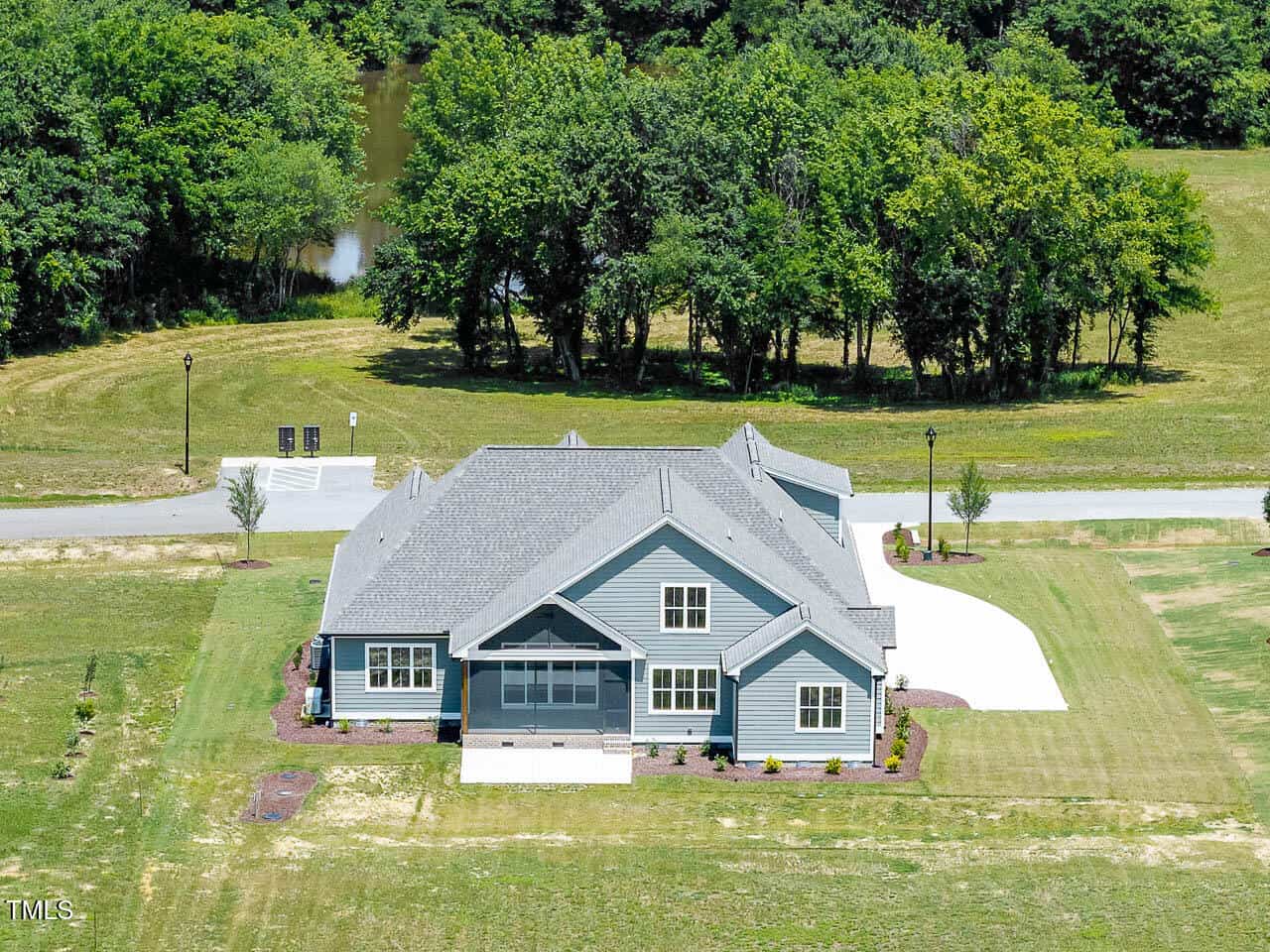 A blue-gray house with white trim sits on a spacious grassy lot. There is a circular driveway, landscaped flower beds, and a wooded area with a pond in the background. The scene is bright and clear.