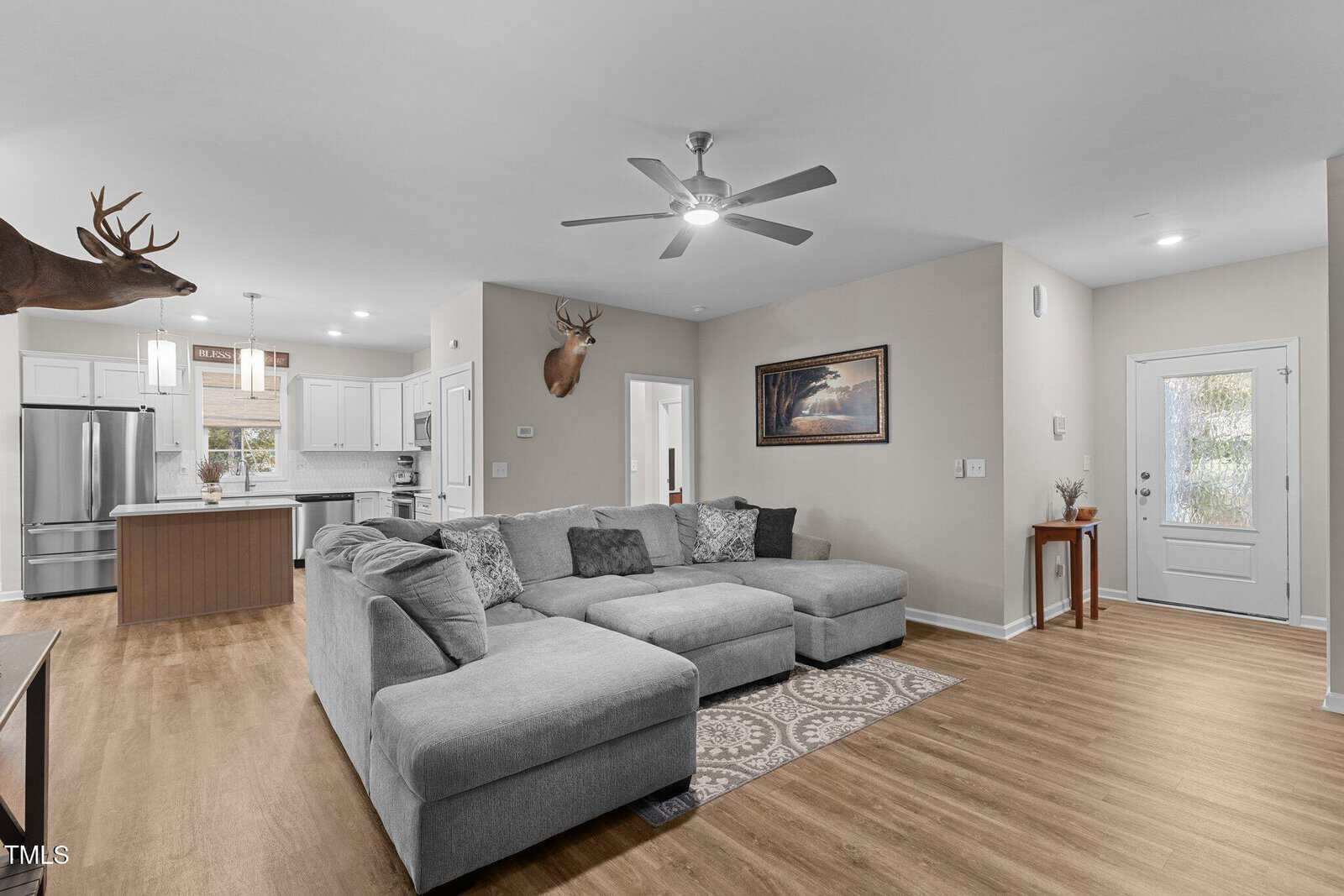 A spacious living room with a large gray sectional sofa on a patterned rug. The room has wooden floors, a ceiling fan, and a wall-mounted deer head. The open kitchen has white cabinets and a central island. A photo hangs on the wall near the entrance.