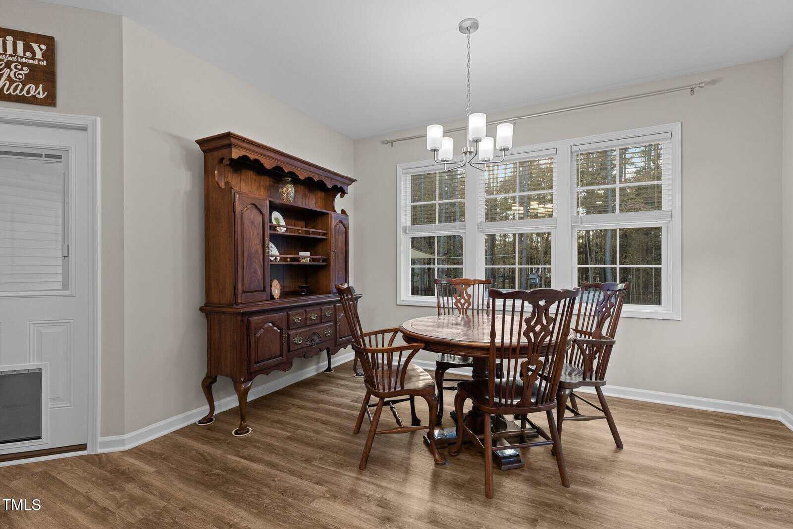 A dining room with a wooden round table and six chairs. A hutch with decorative items is against the left wall, and a window on the right allows natural light. A modern chandelier hangs above the table. The floor is wood, and the walls are light-colored.