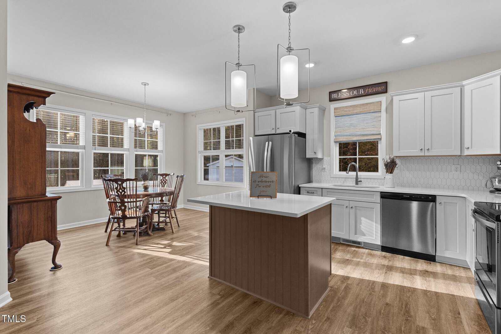 A modern kitchen with white cabinets, stainless steel appliances, and a central island with pendant lights. A wooden dining table with chairs is near large windows. The room has hardwood floors and light-colored walls.
