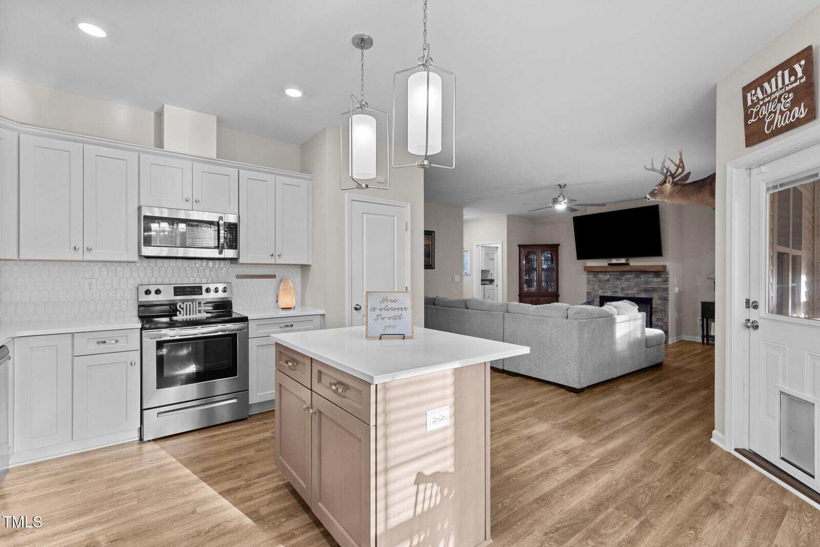 Modern kitchen and living area with wooden floors. The kitchen features white cabinets, a stainless steel stove, and a central island with pendant lights. The living area has a gray sectional sofa, a wall-mounted TV, and a deer head mounted above a fireplace.