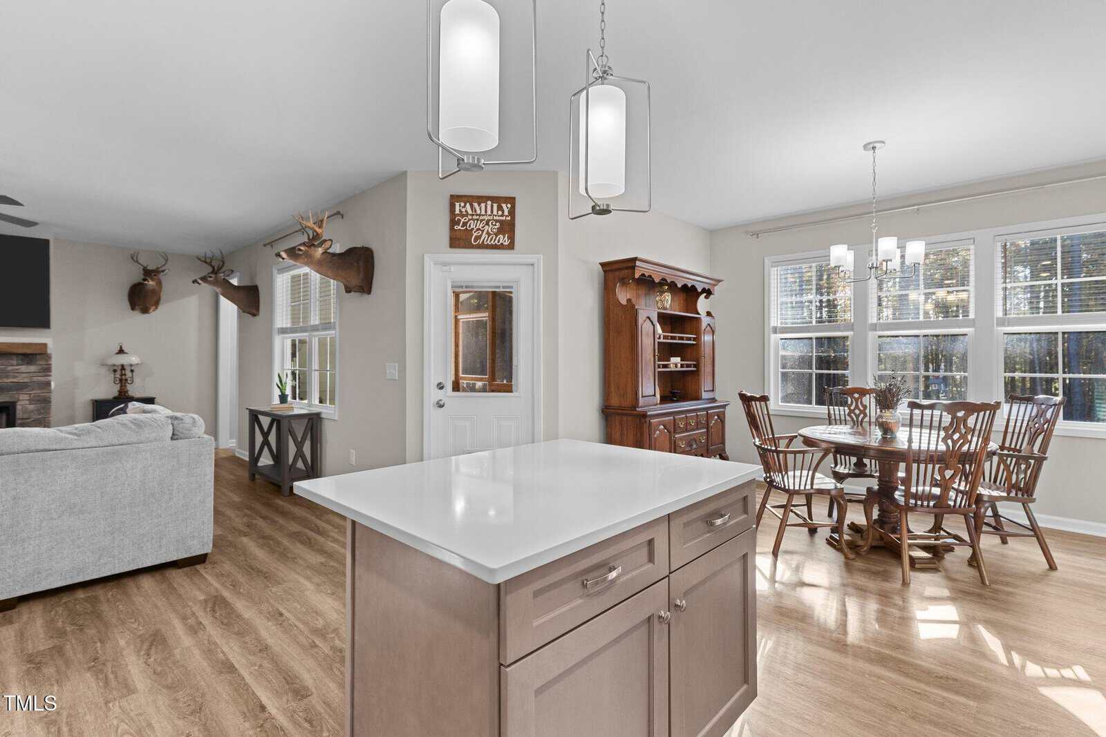 A modern open-concept kitchen and dining area with light wood flooring. The kitchen features a sleek island with two pendant lights above. A dining table is placed near large windows, and mounted deer heads are visible on the walls in the living area.