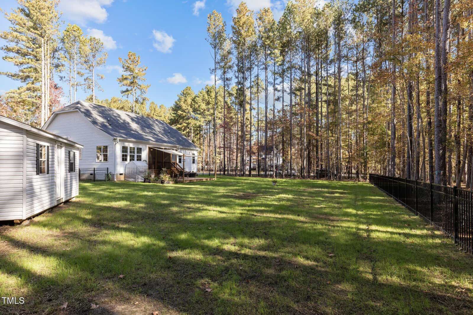 A single-story white house with a porch is surrounded by a grassy yard. A black fence borders one side. Trees with autumn leaves line the backyard, and a small shed is visible on the left. The sky is clear with a few clouds.