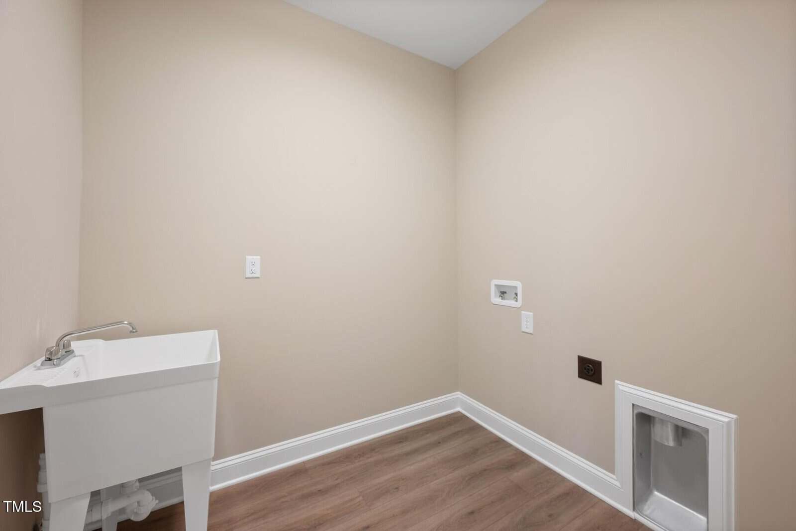A laundry room with beige walls, wood-style flooring, a white utility sink on the left, and hookups for a washer and dryer on the far wall. There is also a pet door in the wall on the right.