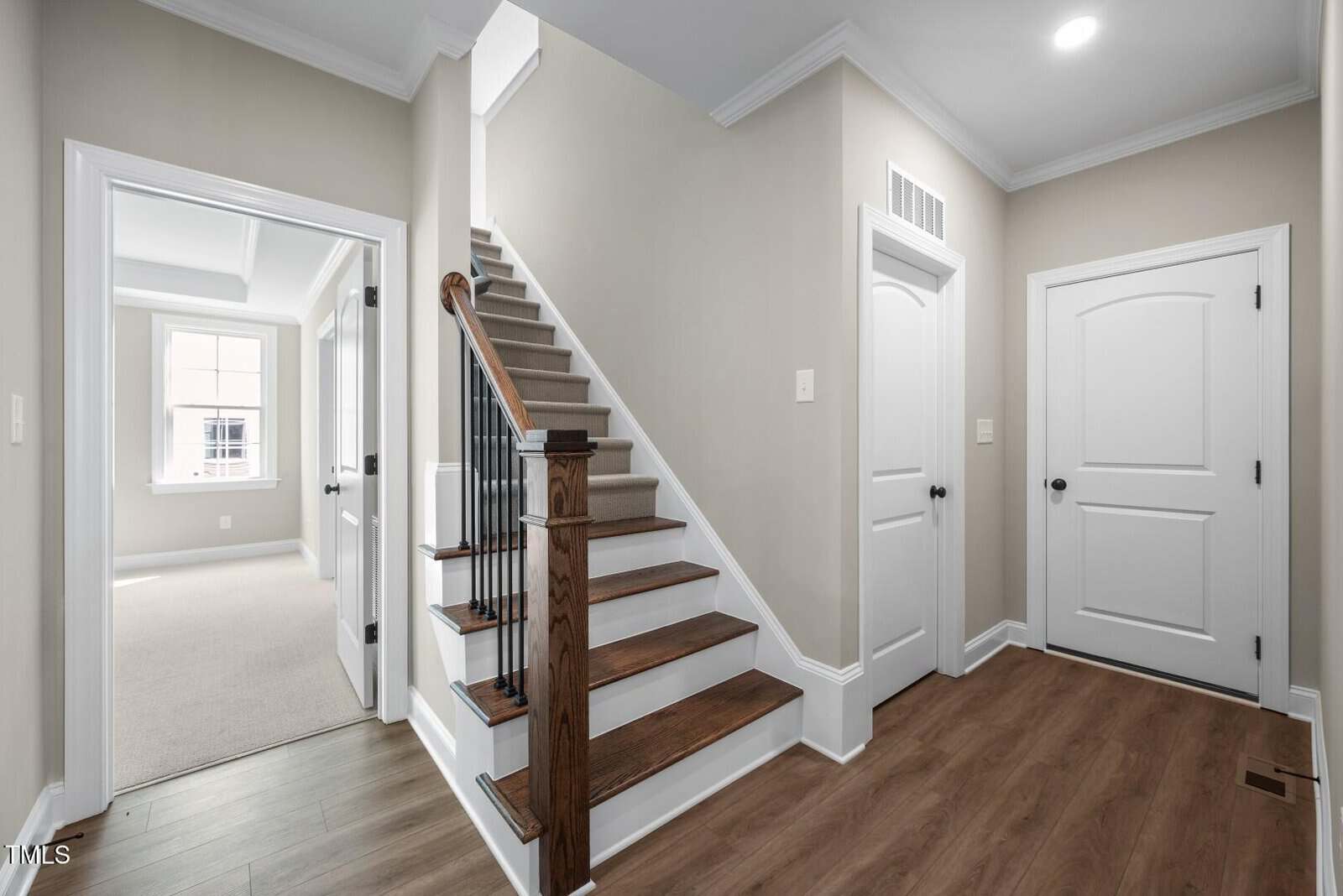 A hallway with wood flooring, beige walls, and white trim. A staircase with a wooden handrail and carpeted steps leads upward. Three white doors are visible, one open to a bright room with a window.