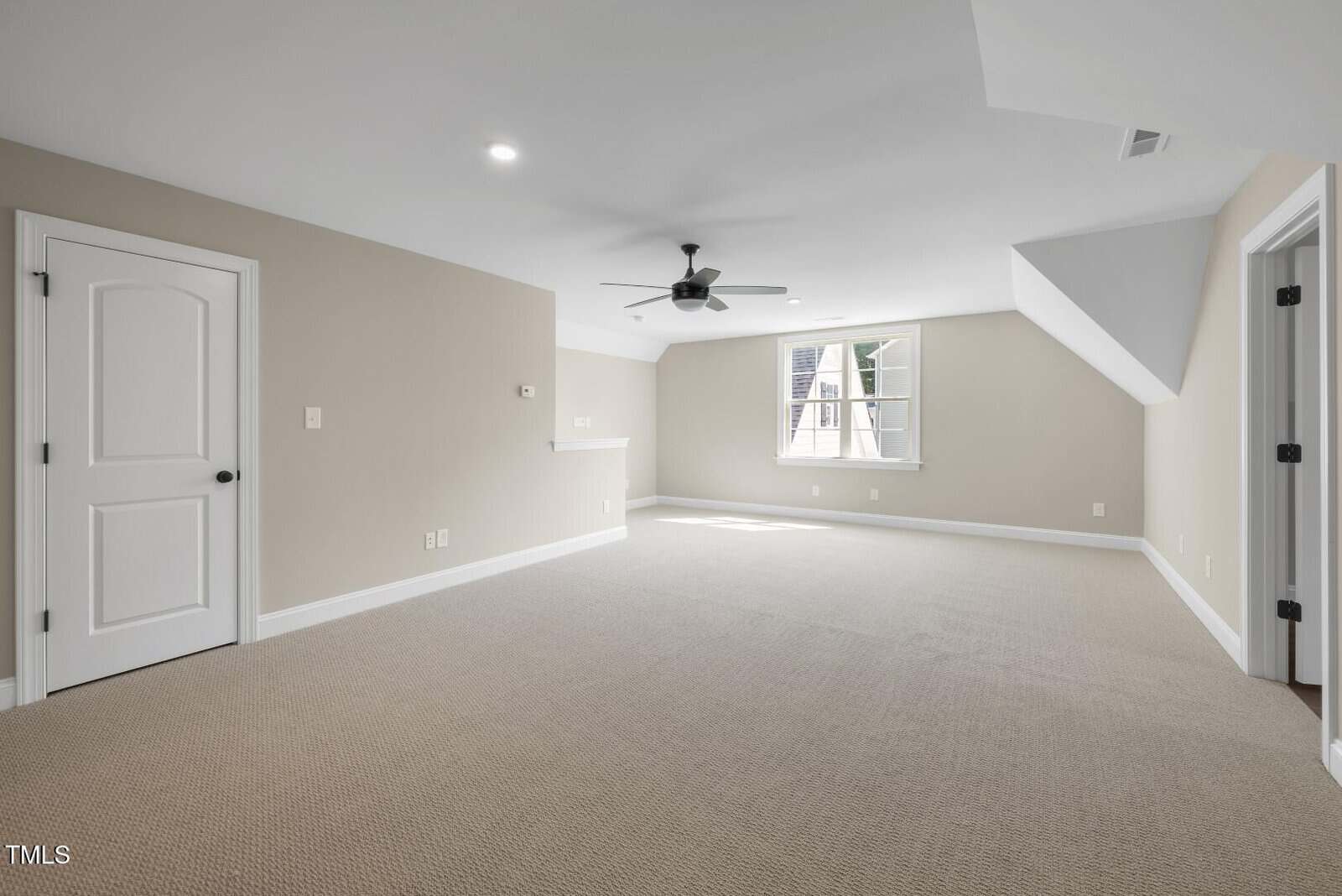 Spacious, empty carpeted room with beige walls, white trim, a ceiling fan, recessed lighting, and a window letting in natural light. There are two white doors and an alcove area on one side.