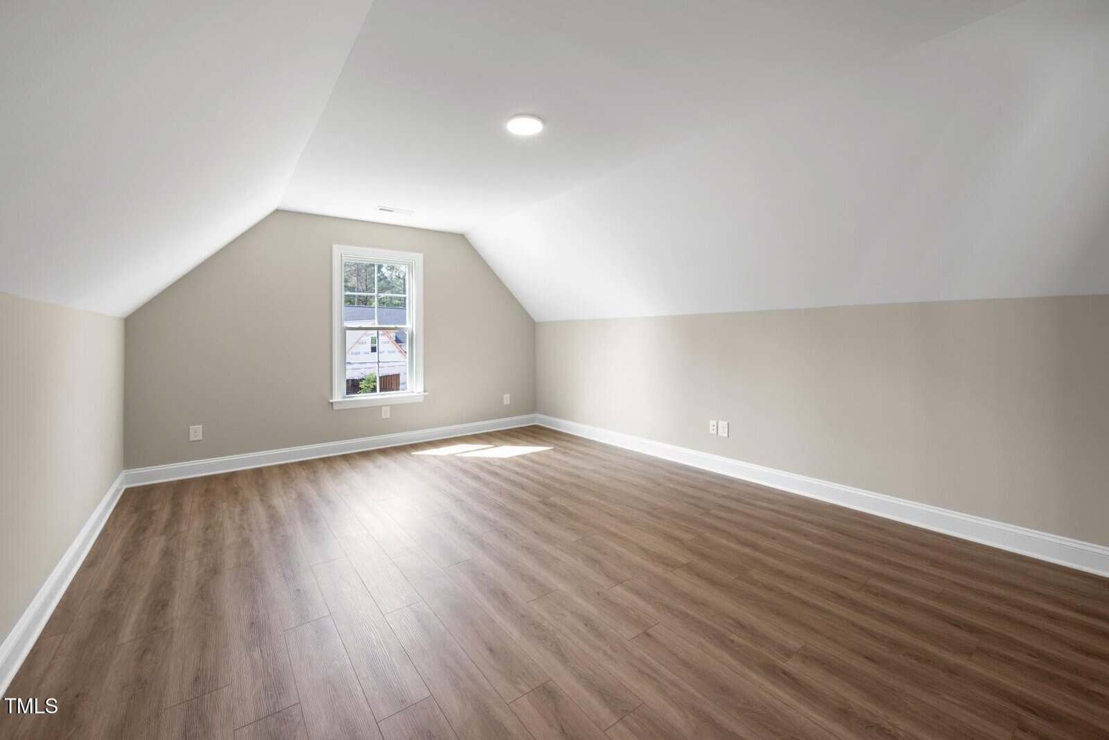 An empty attic room with sloped ceilings, beige walls, wood flooring, a small window letting in sunlight, and a recessed ceiling light.