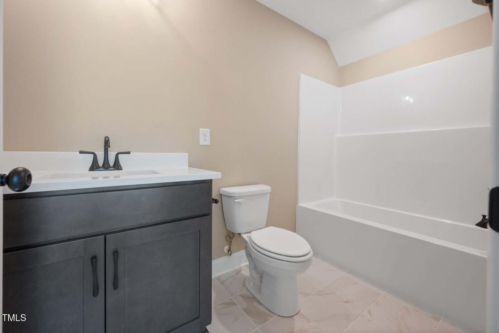 A bathroom with beige walls, a white bathtub-shower combination, a white toilet, and a dark vanity with a white countertop and black faucet. The floor is tiled in a light color.