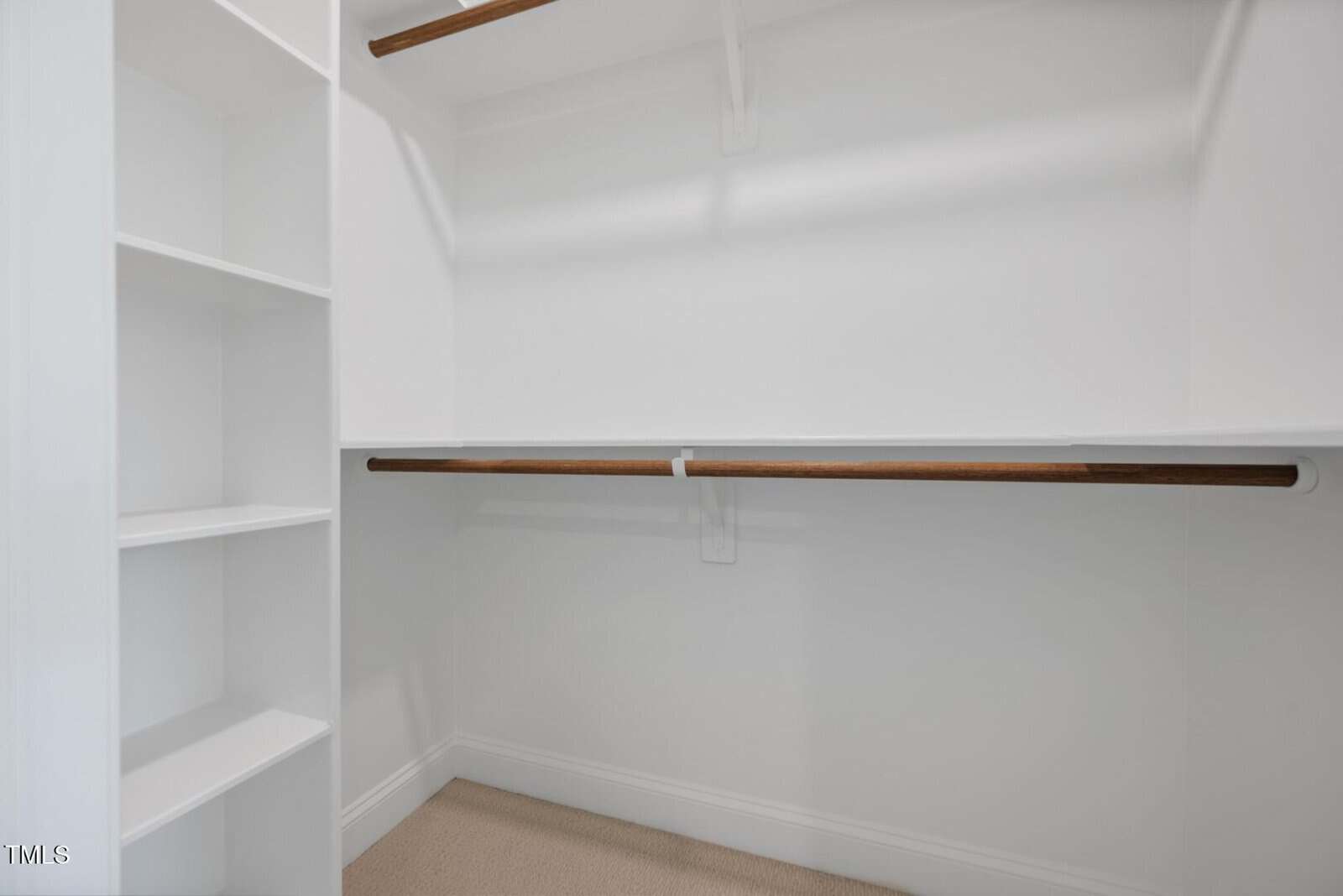 Empty white walk-in closet with built-in shelves on the left and two wooden hanging rods along the right and back walls. The floor is carpeted in a light beige color.