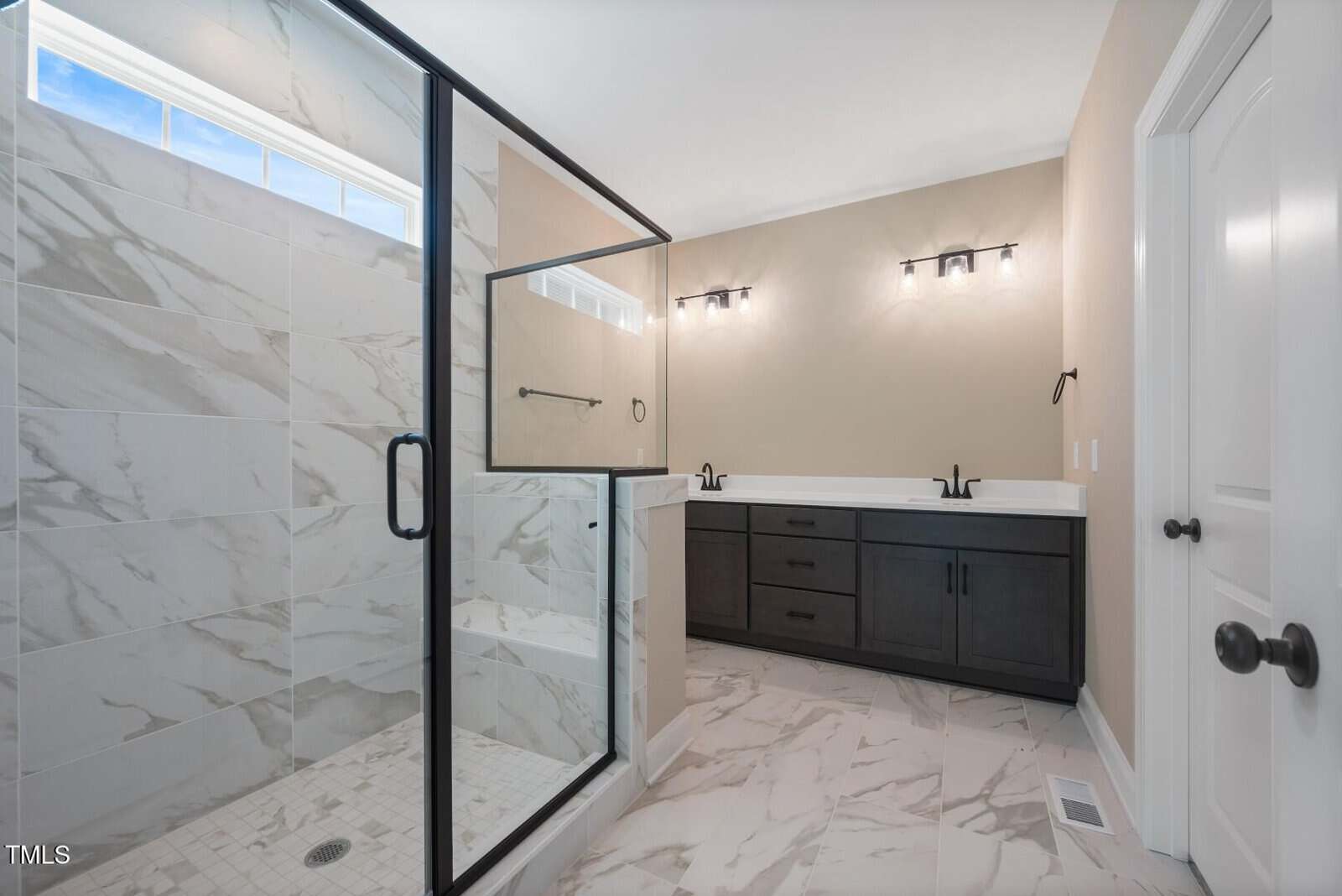 Modern bathroom with a glass shower, marble tile floors and walls, double-sink vanity with black cabinets, and two wall-mounted light fixtures above the mirrors. Neutral wall color and natural light from a high window.