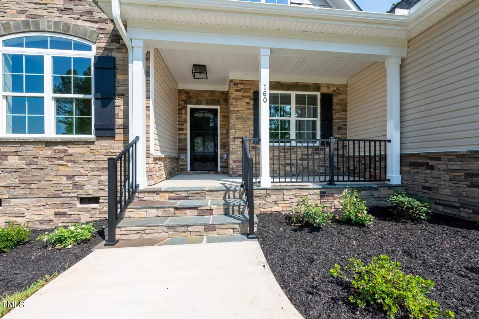 Front view of a house entry with stone and siding exterior, black metal railings on both sides of the steps, a covered porch, a built-in planter with shrubs, and a large window to the left of the front door.