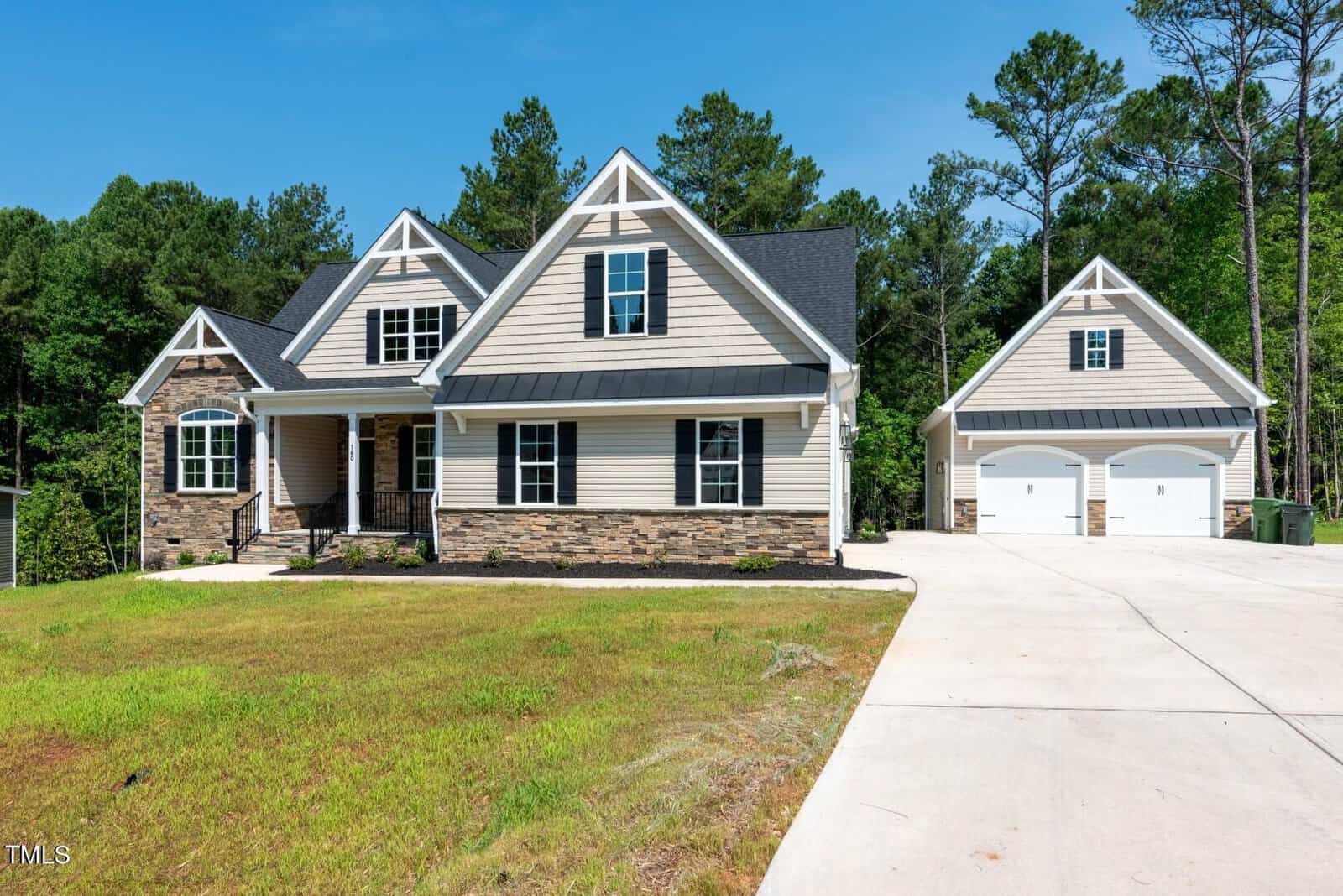 A beige and stone two-story suburban house with gable roofs, a front porch, and an attached garage. A separate two-car garage is on the right. The house sits on a large driveway with a grassy front yard and trees in the background.
