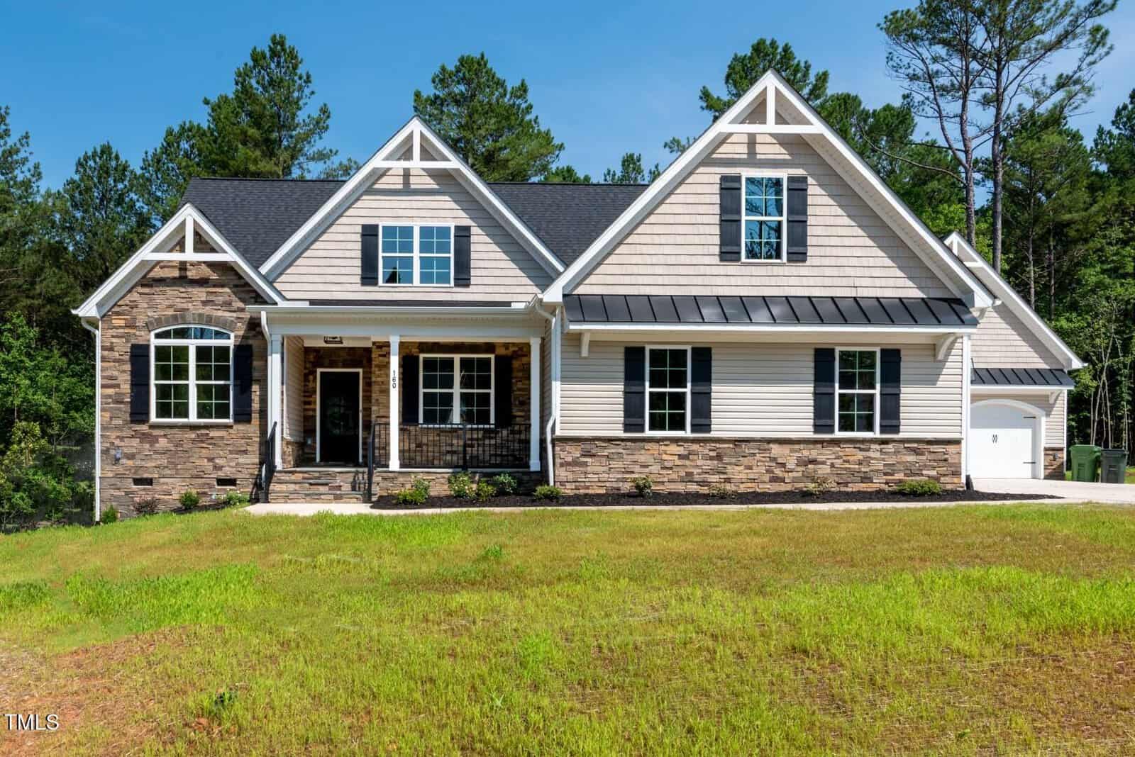 A modern two-story suburban house with light-colored siding, stone accents, black roof, covered front porch, and an attached garage, set on a grassy lawn with trees in the background.