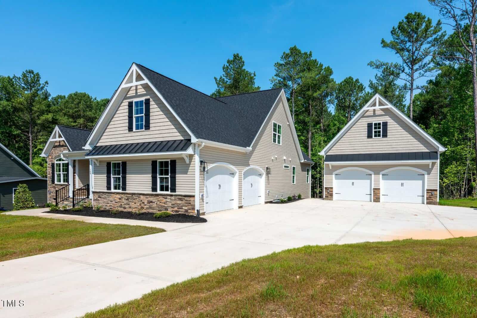 A modern two-story house with beige siding, dark shingles, and stone accents, featuring a double garage attached to the house and a separate double garage, both set on a wide concrete driveway with green lawn and trees in the background.