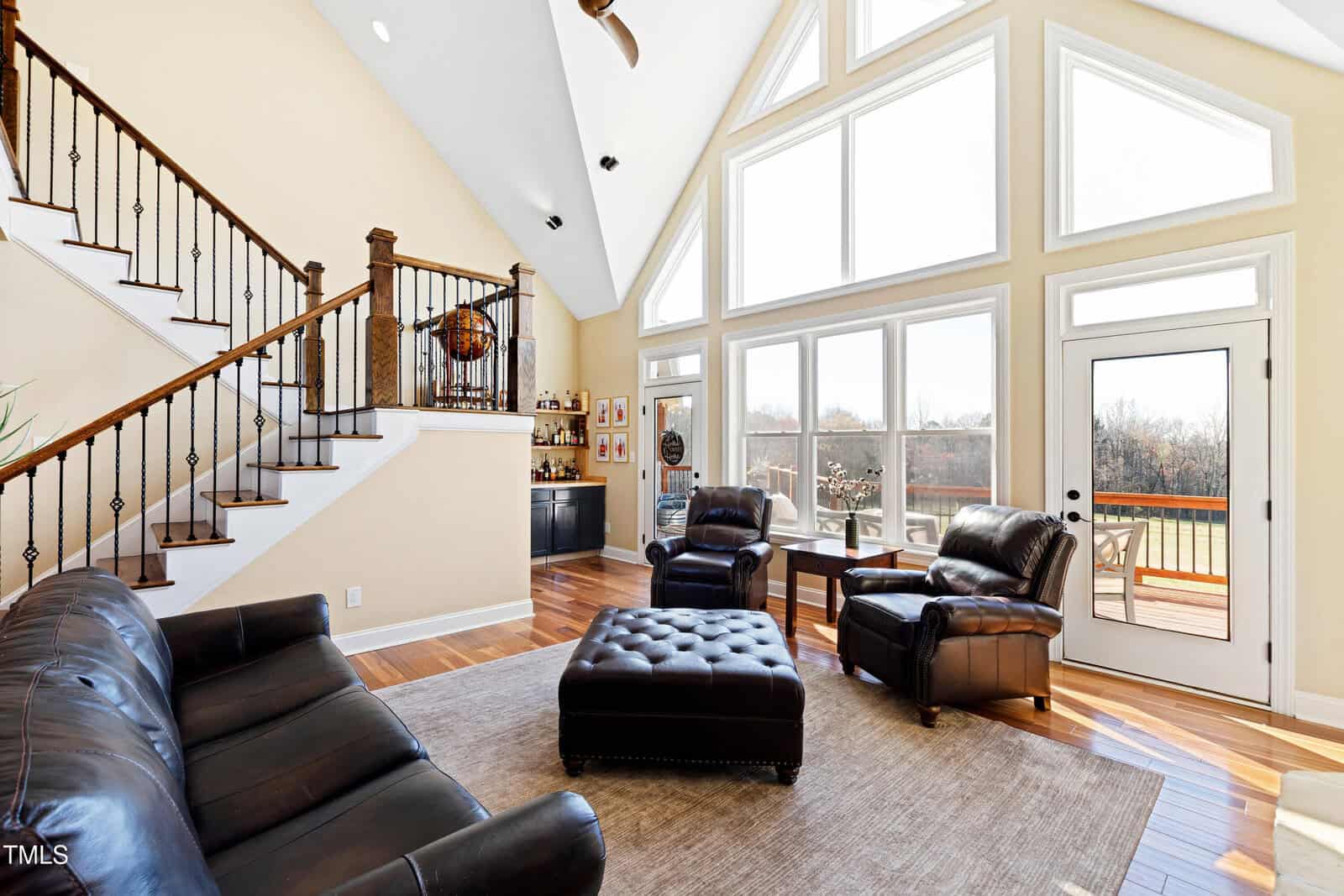 Spacious living room with large windows and high ceilings. It features leather sofas, a central ottoman, and a view of the outdoor deck. A staircase with a decorative railing leads upstairs, and a small bar area is visible to the side.