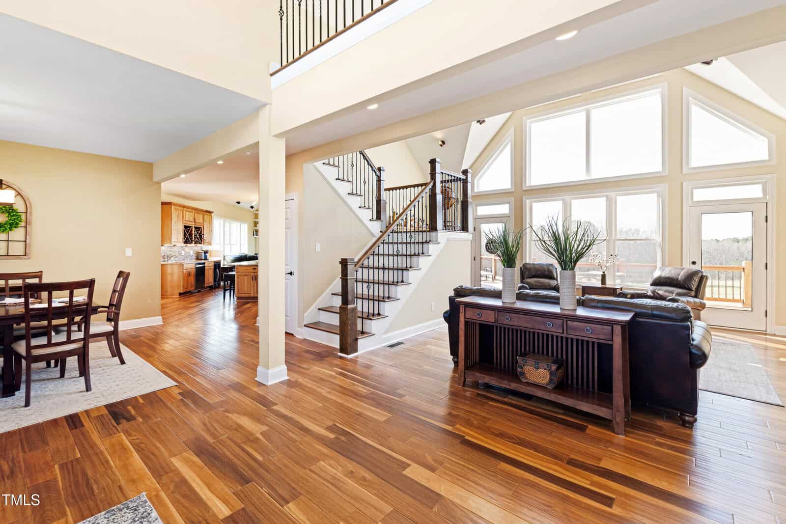 Spacious living area with hardwood floors, large windows, and a high ceiling. A staircase with a dark wood railing leads to an upper level. The room features leather sofas, a console table, and a view into a dining and kitchen area.
