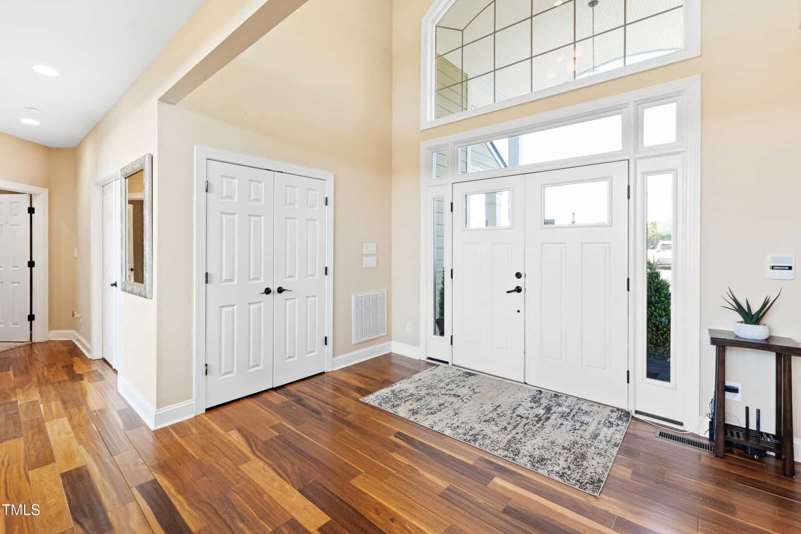 A spacious entryway with light-colored walls and hardwood flooring. There are double front doors with large windows above, allowing natural light in. A patterned rug lies on the floor, and a double-door closet is on the left.
