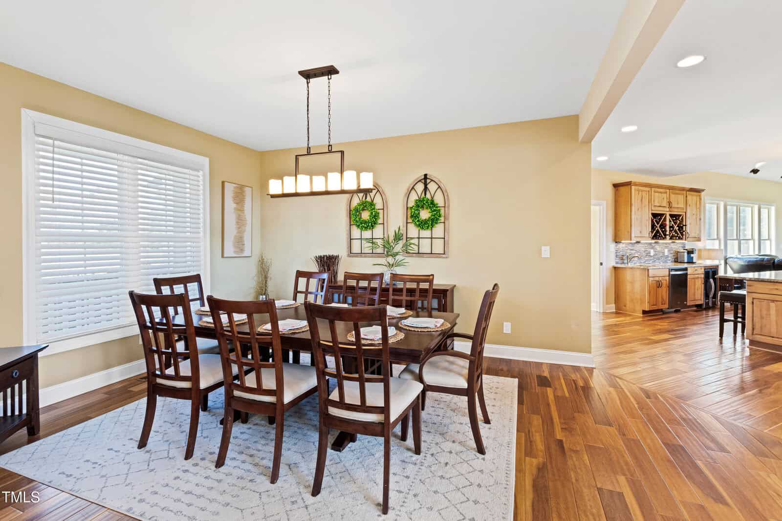 A dining room with a dark wooden table and eight chairs on a patterned rug. A modern chandelier hangs above. Two decorative pieces with green wreaths are on the wall. Large window with blinds on the left. Kitchen visible in the background.