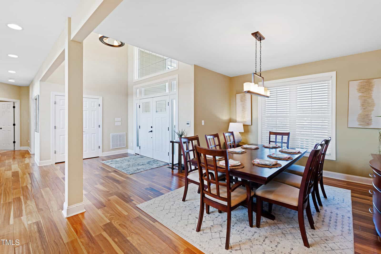 A dining room with a wooden table and eight chairs, set with placemats. A modern rectangular chandelier hangs above. The room has hardwood floors, beige walls, and a large window with blinds. A hallway and entrance with double doors are visible.