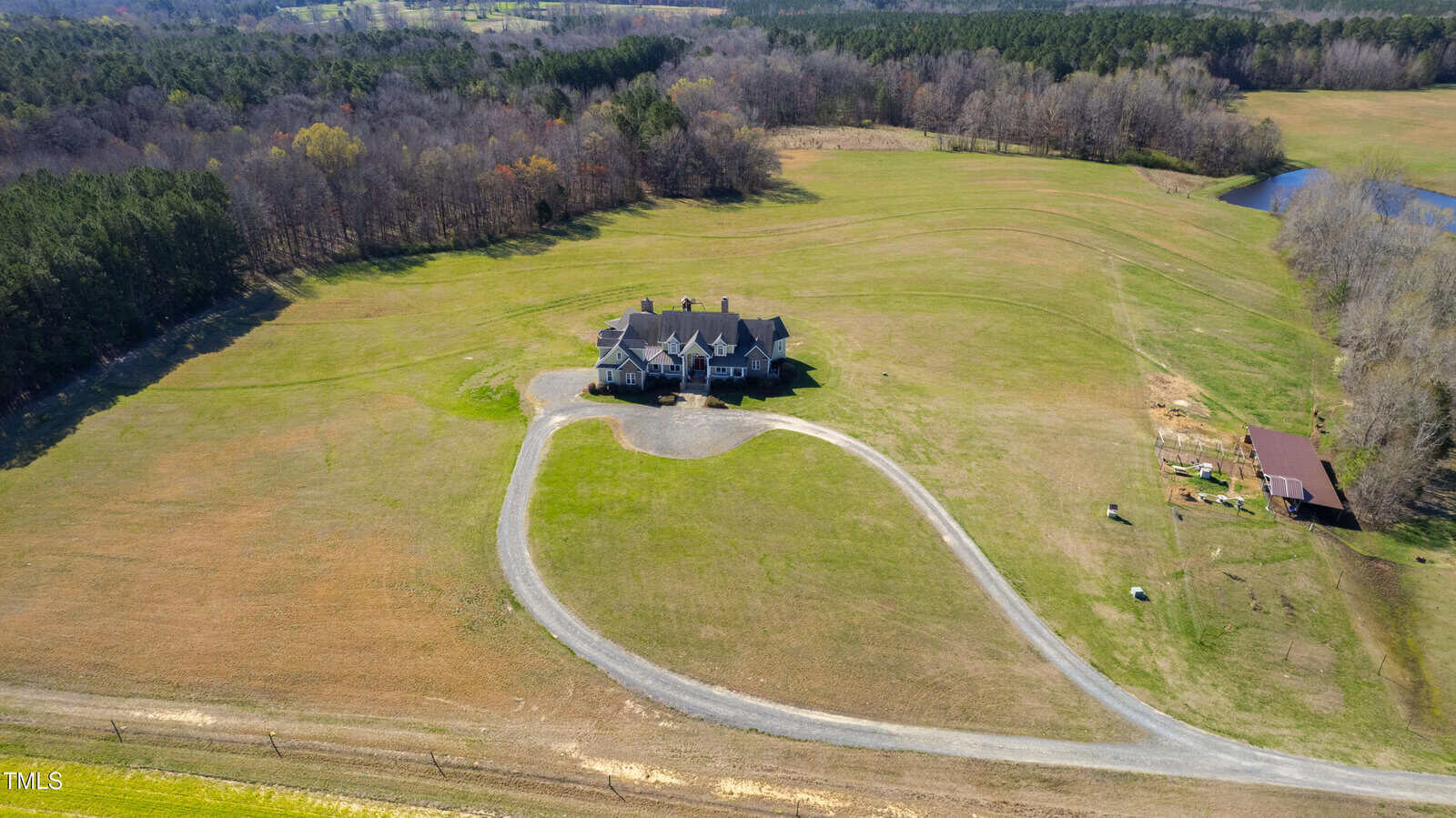 Aerial view of a large house surrounded by expansive grassy fields. A winding driveway leads to the house. Nearby, there is a small pond and wooded areas in the background.