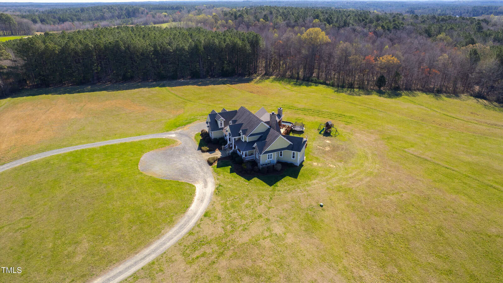 Aerial view of a large, multi-story house with a dark roof, situated in a vast, open field. A gravel driveway leads to the house. Surrounding the property are expansive lawns and a dense line of trees in the background.
