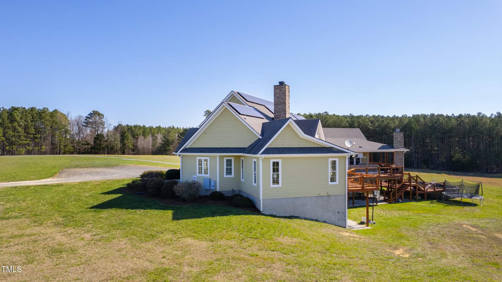 A yellow house with a large wooden deck and chimney is set in a grassy area surrounded by trees under a clear blue sky. The driveway leads up to the house, and a trampoline is visible next to the deck.