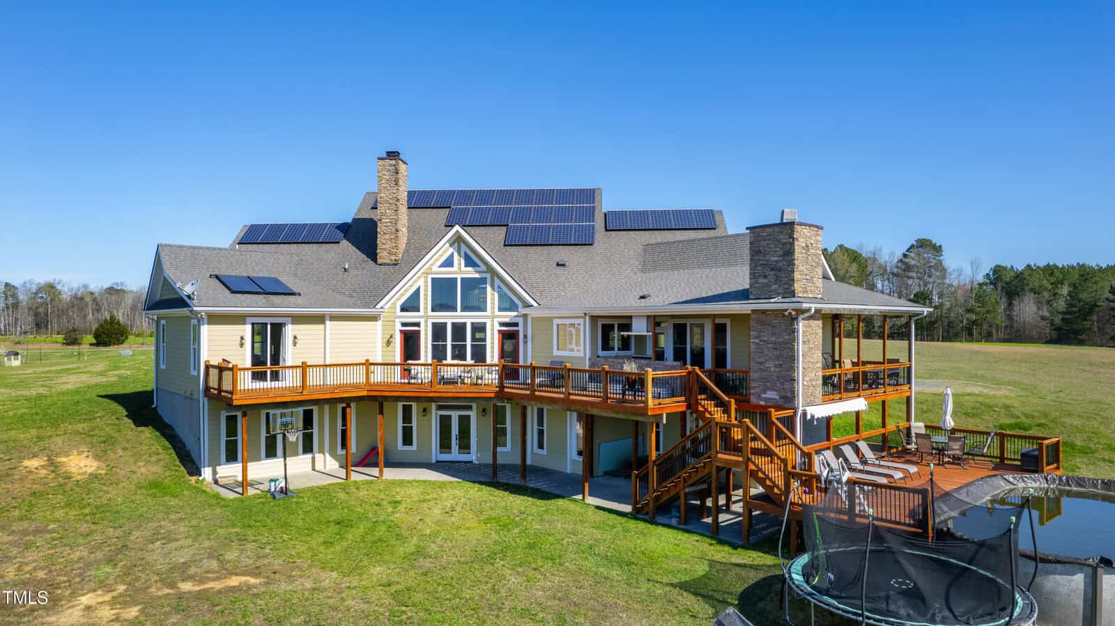 A large, modern house with solar panels on the roof, featuring a wraparound wooden deck and multiple windows. There's a trampoline and a round pool in the spacious backyard, surrounded by open grassy fields and trees under a clear blue sky.