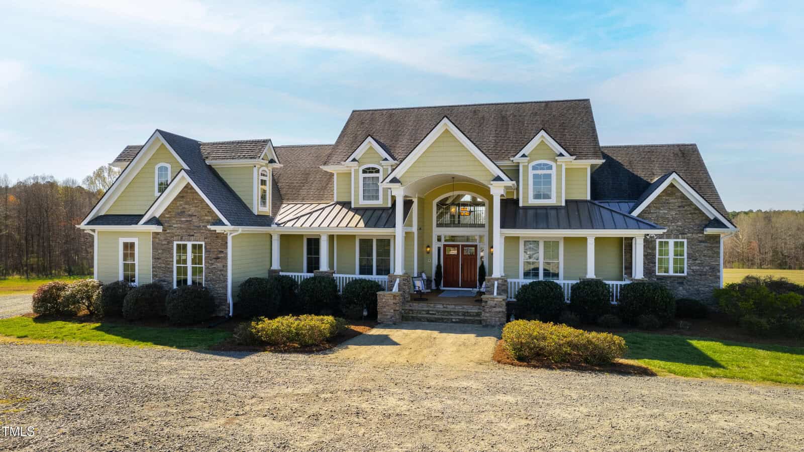 Nestled on 3220 Bliss Trail, this large two-story house with beige siding and stone accents sits on a spacious property. It features multiple peaked roofs, a red front door with a white archway, and a gravel driveway leading to the entrance. Shrubs line the front yard.