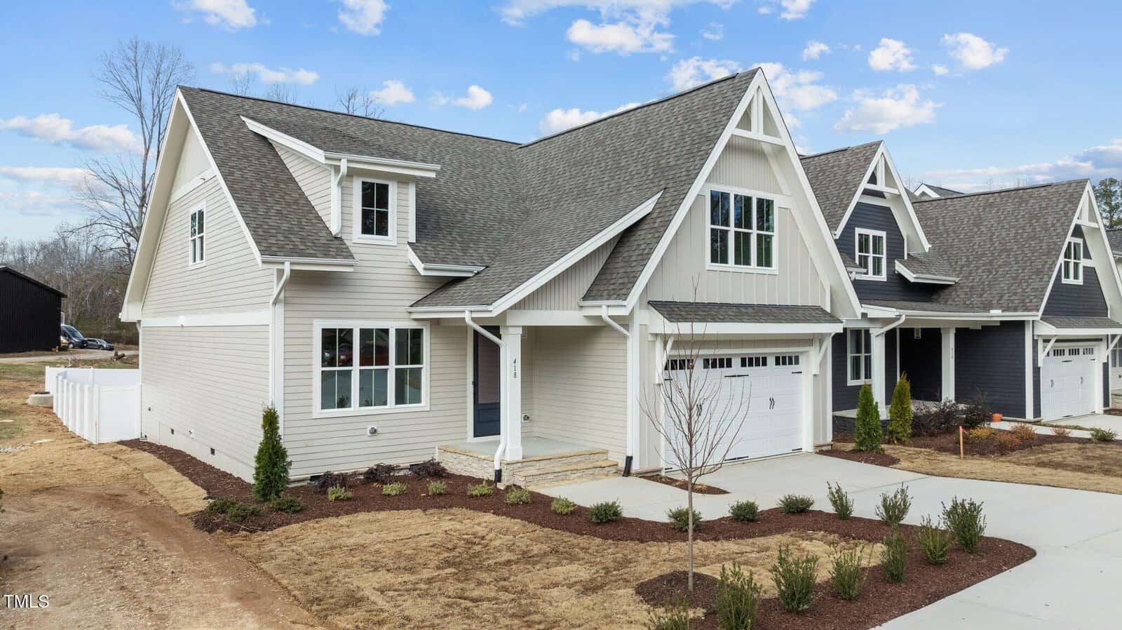 A light gray, two-story suburban house with white trim, a gabled roof, double garage, and a small front yard with new landscaping. Neighboring homes and a partially fenced backyard are visible. Sky is partly cloudy.