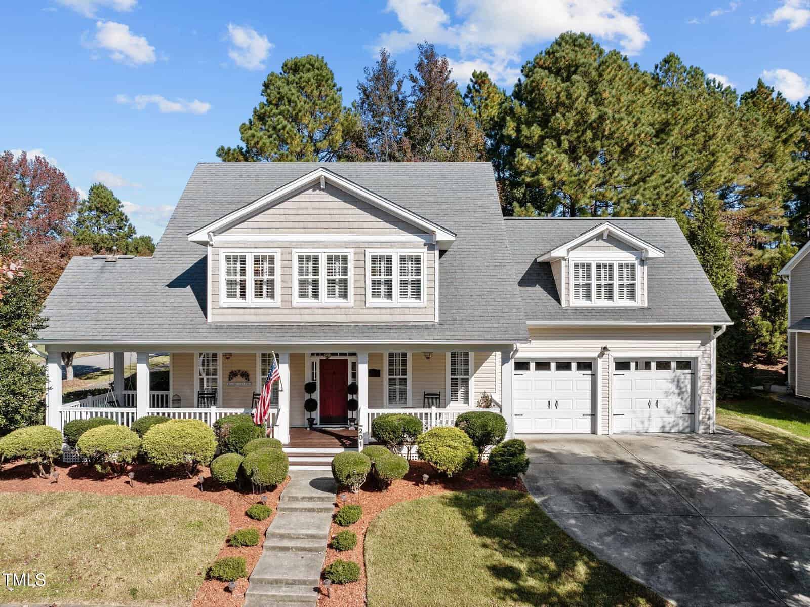 Located at 201 Oxford Park Boulevard, this charming two-story gray house features a welcoming front porch and is embraced by neatly trimmed bushes. A driveway leads to the double garage, while tall trees stand in the background. Near the entrance, an American flag proudly waves under a gentle sky.
