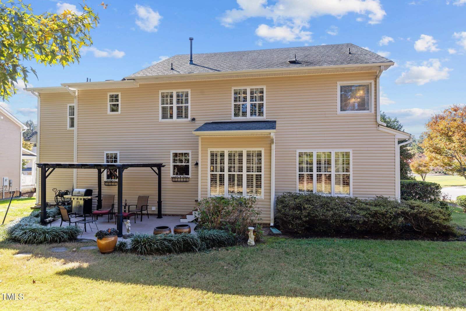 A two-story beige house with multiple windows and a small covered patio in the backyard. The patio has a pergola and outdoor furniture. The scene includes some shrubbery and lawn, under a partly cloudy sky.