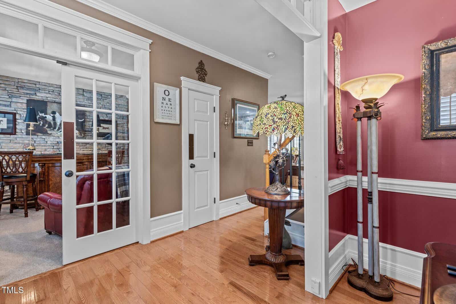 An interior view of a home with wood flooring. On the right, a stained glass lamp and a tall, decorative lamp are next to a red-painted wall. A doorway with glass panels, leading to another room with stone walls and a wooden table, is visible.