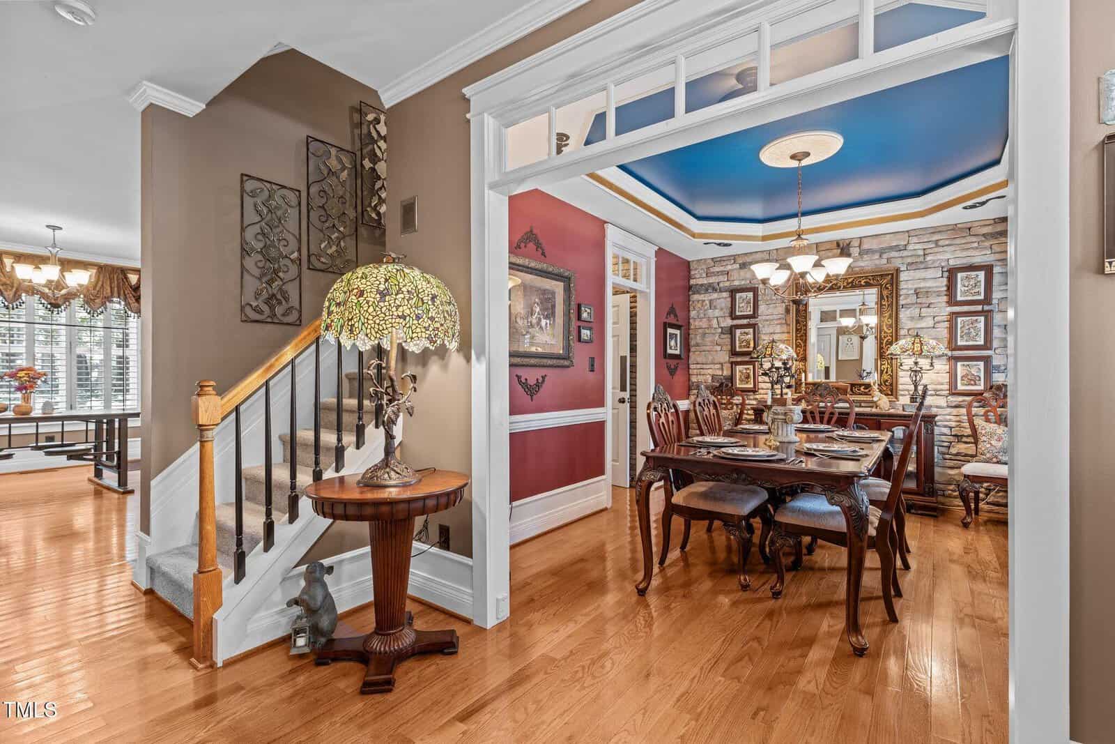 A dining room with a wooden table set for six and a decorative chandelier above. The adjacent hallway features a staircase with a wooden banister and a side table with a stained-glass lamp. Walls are adorned with framed artwork.
