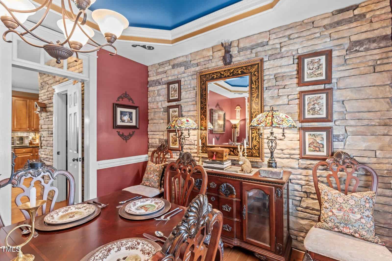 Dining room with a stone accent wall, featuring a large mirror and decorative frames. A wooden table is set with plates and silverware. Decorative chairs surround the table. A chandelier hangs above, and ornate lamps are placed on a wooden sideboard.