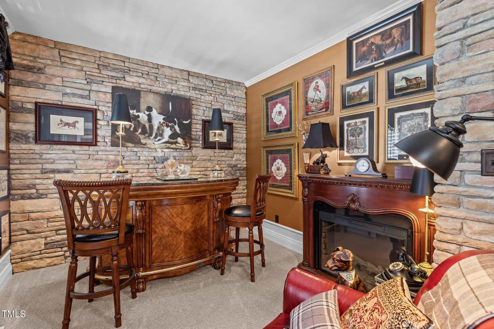 A cozy room with a stone accent wall features a wooden bar with two high chairs. Various art pieces and photos adorn the walls. A decorative fireplace sits nearby, with table lamps and a red sofa adding warmth to the space.