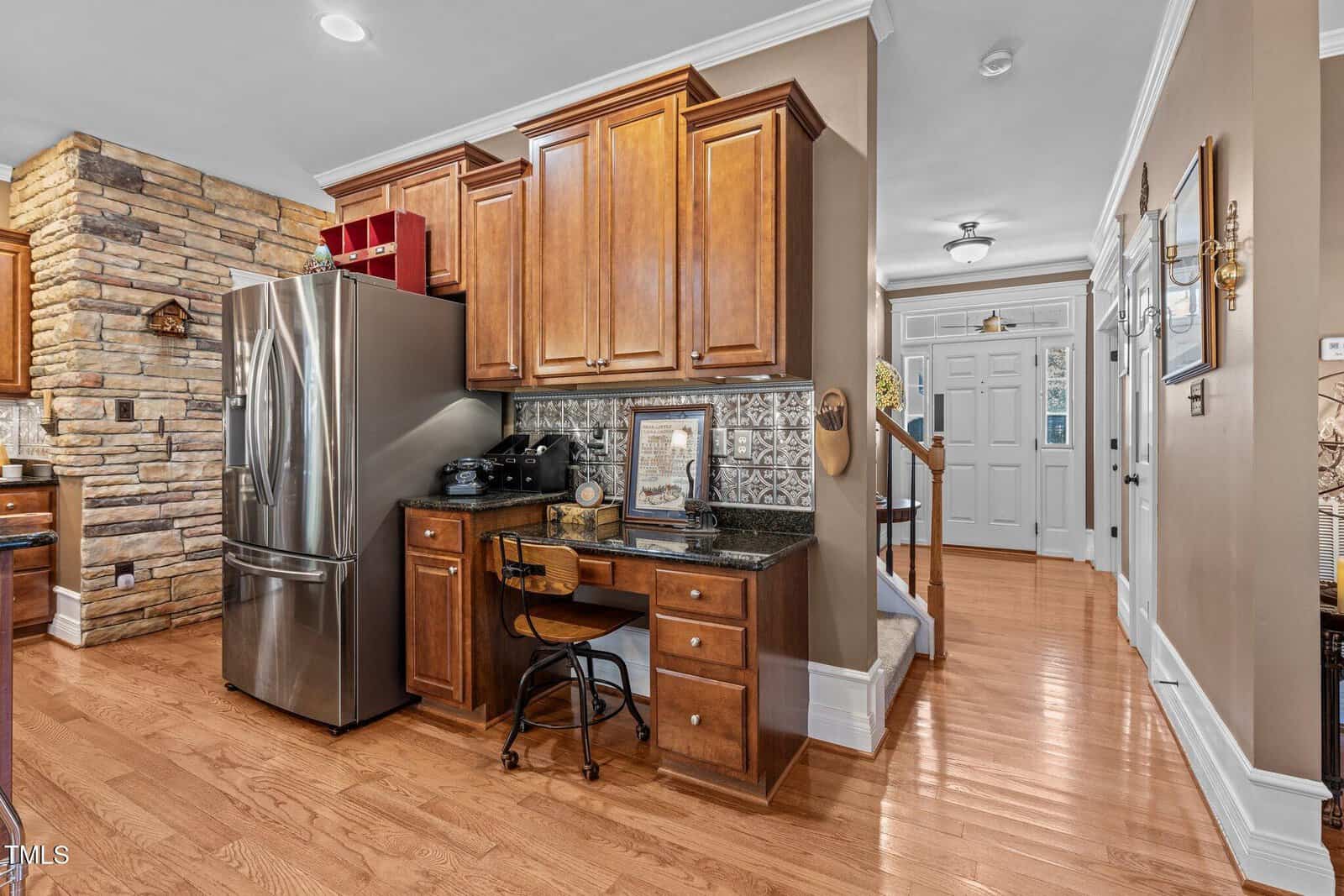 A kitchen features wooden cabinets, a stainless steel refrigerator, and a small desk with a chair. The desk area includes a framed artwork, a lamp, and a coffee maker. The room has hardwood flooring, light-colored walls, and a stone accent wall.