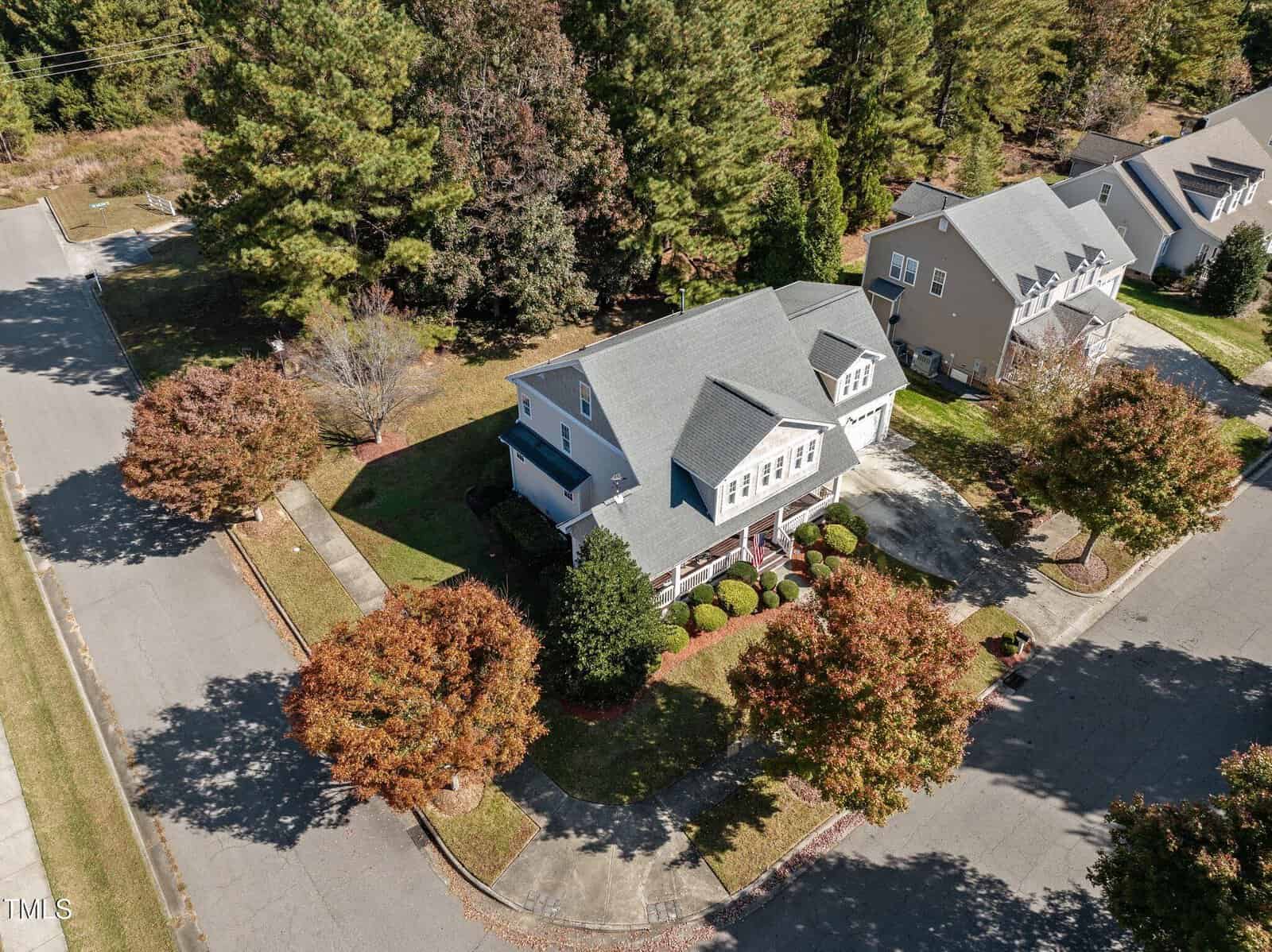 Aerial view of a residential neighborhood featuring a two-story house with a green roof and white siding, surrounded by neatly trimmed bushes and young trees with autumn foliage. The house is situated on a corner lot with adjacent homes nearby.