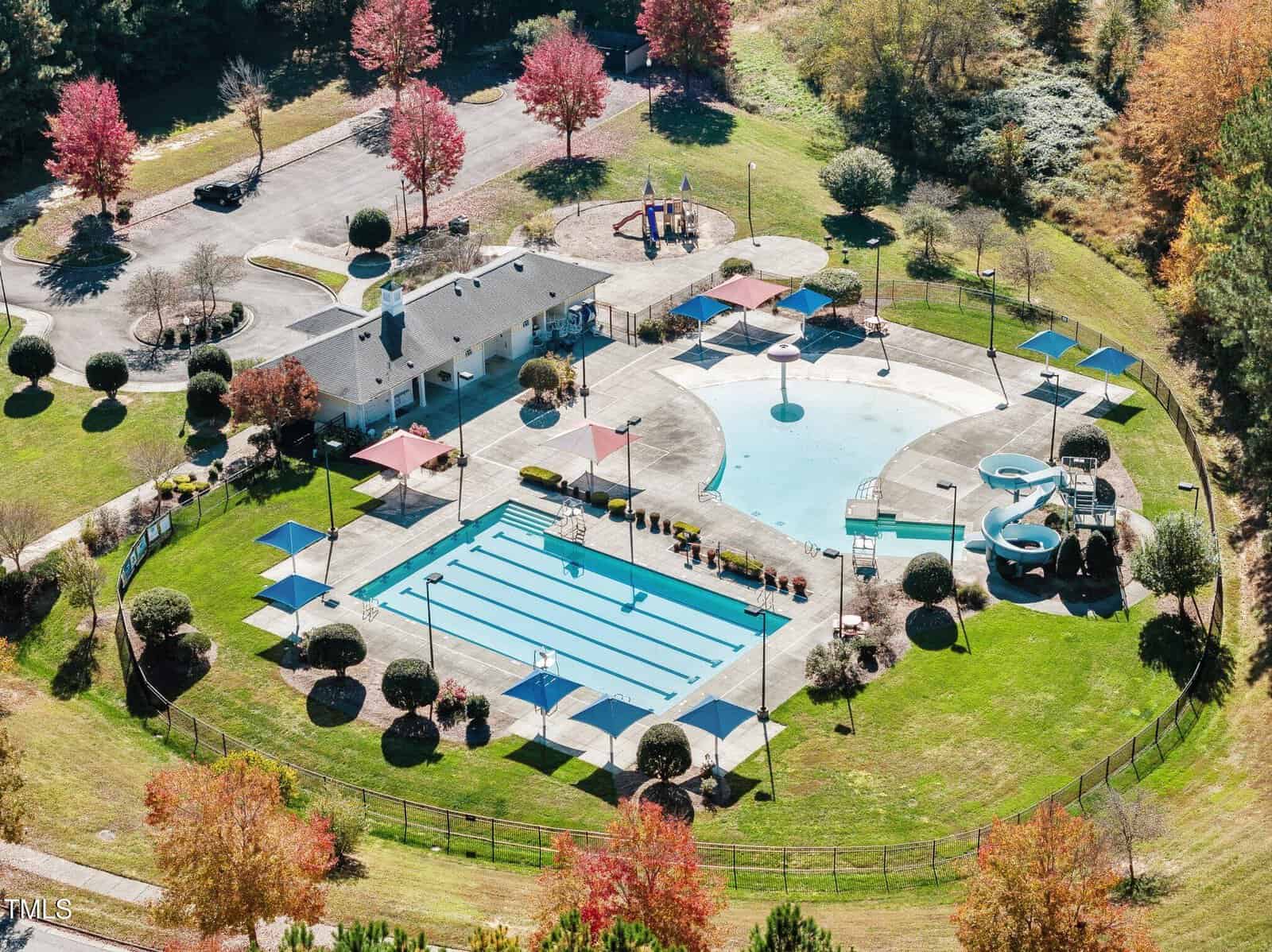Aerial view of a recreational area featuring a large swimming pool with lanes, a children's splash pad, a waterslide, and surrounding grassy areas with picnic tables. The setting includes autumn trees and a nearby parking lot.