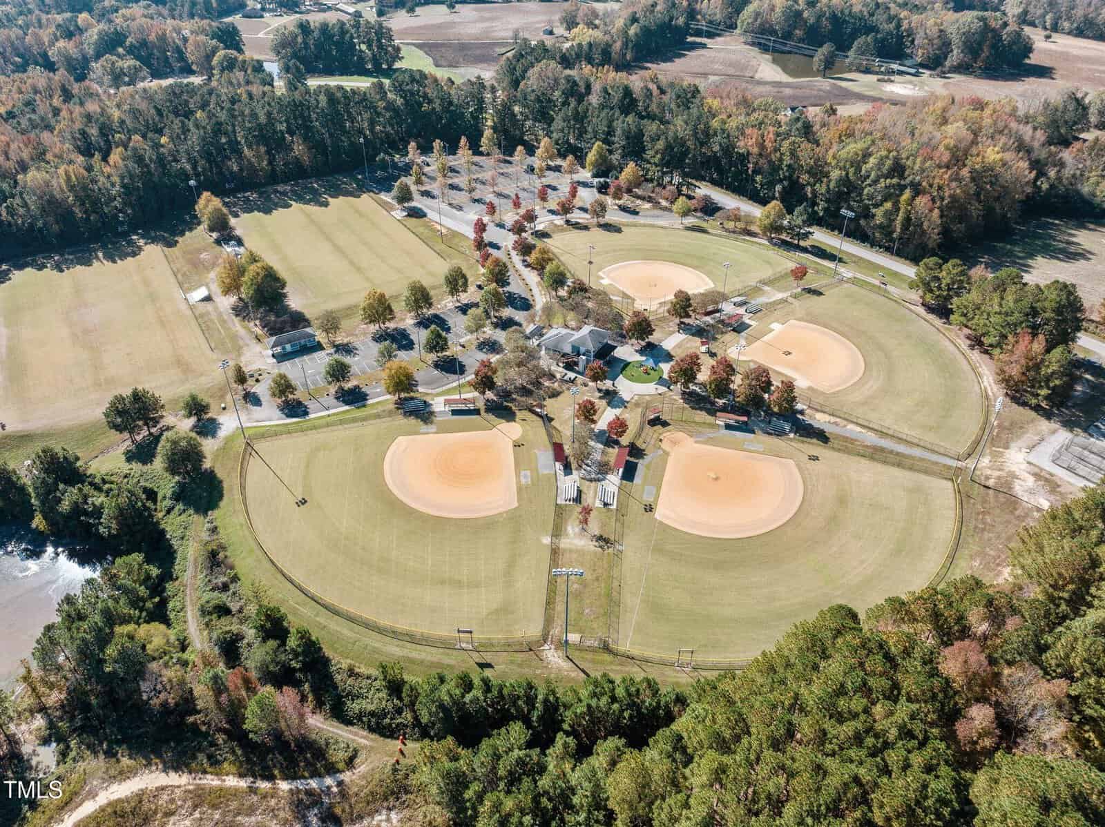 An aerial view of a sports complex featuring multiple baseball and softball fields surrounded by trees. There are five fields forming a star shape, with open grassy areas and a parking lot nearby. Autumn foliage is visible in the surrounding forest.