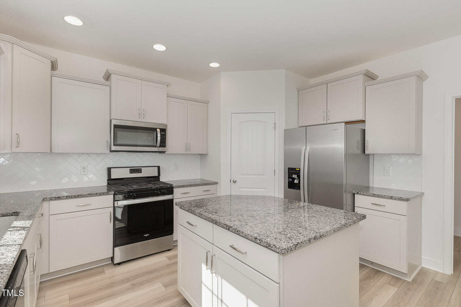 A modern kitchen with white cabinets, stainless steel appliances including a refrigerator, microwave, and stove, granite countertops, a large island, and light wood flooring. Recessed lighting illuminates the space.