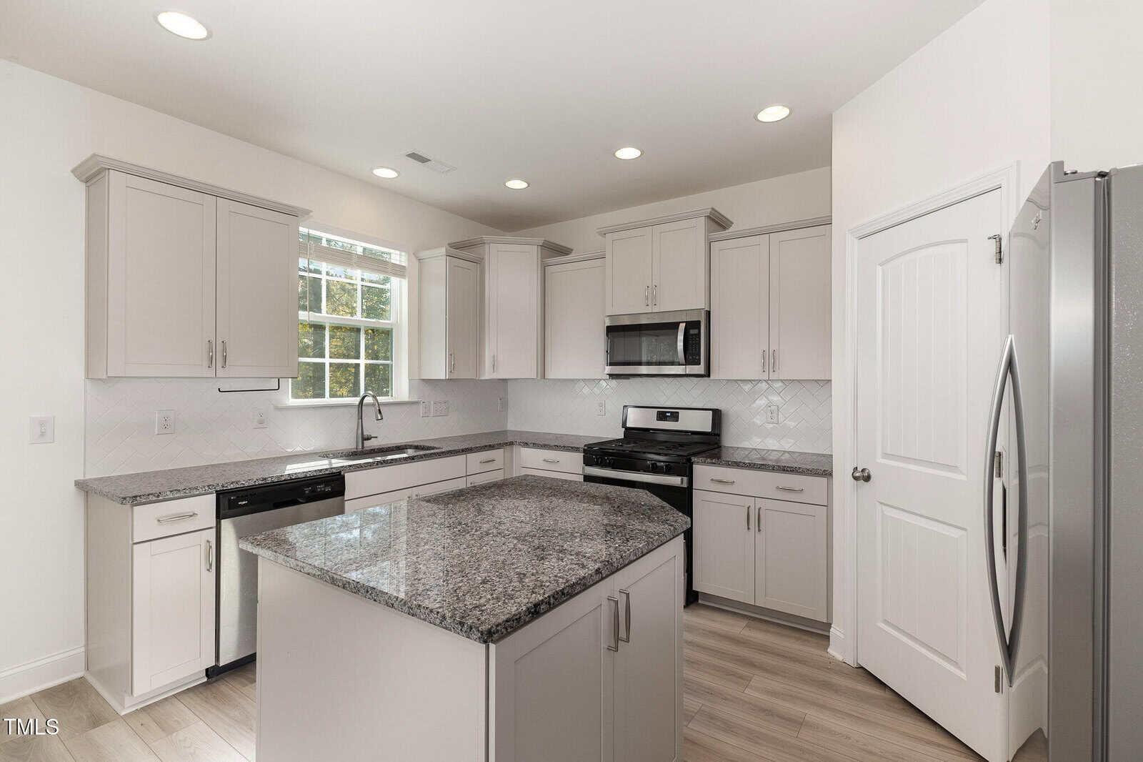 Modern kitchen with gray cabinets, stainless steel appliances, granite countertops, center island, window over the sink, and light wood flooring. Recessed lighting brightens the space.