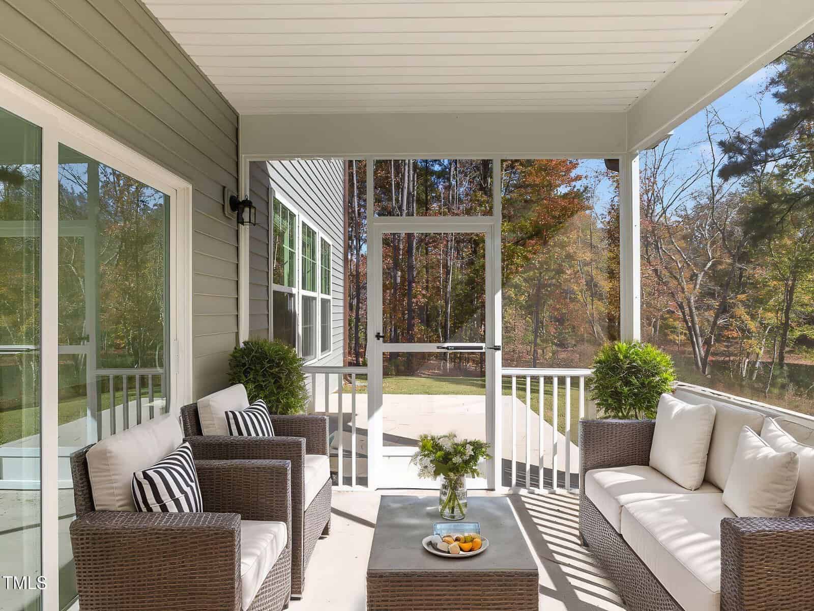 Screened-in porch with wicker furniture, including a sofa and two armchairs with striped pillows, a coffee table with a vase and snacks, potted plants, and views of a backyard with trees in autumn.