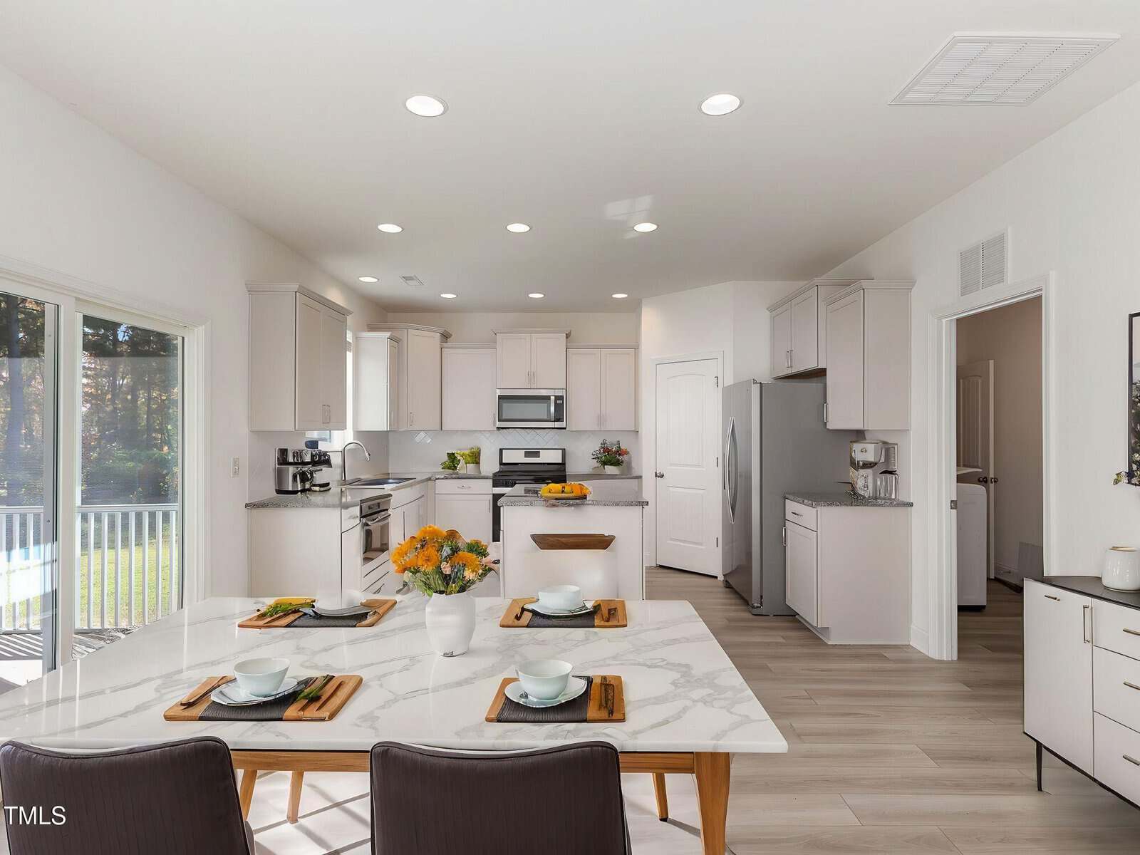 Modern, bright kitchen with white cabinets, stainless steel appliances, and a marble dining table set for four, decorated with orange flowers. Large sliding glass doors let in natural light.