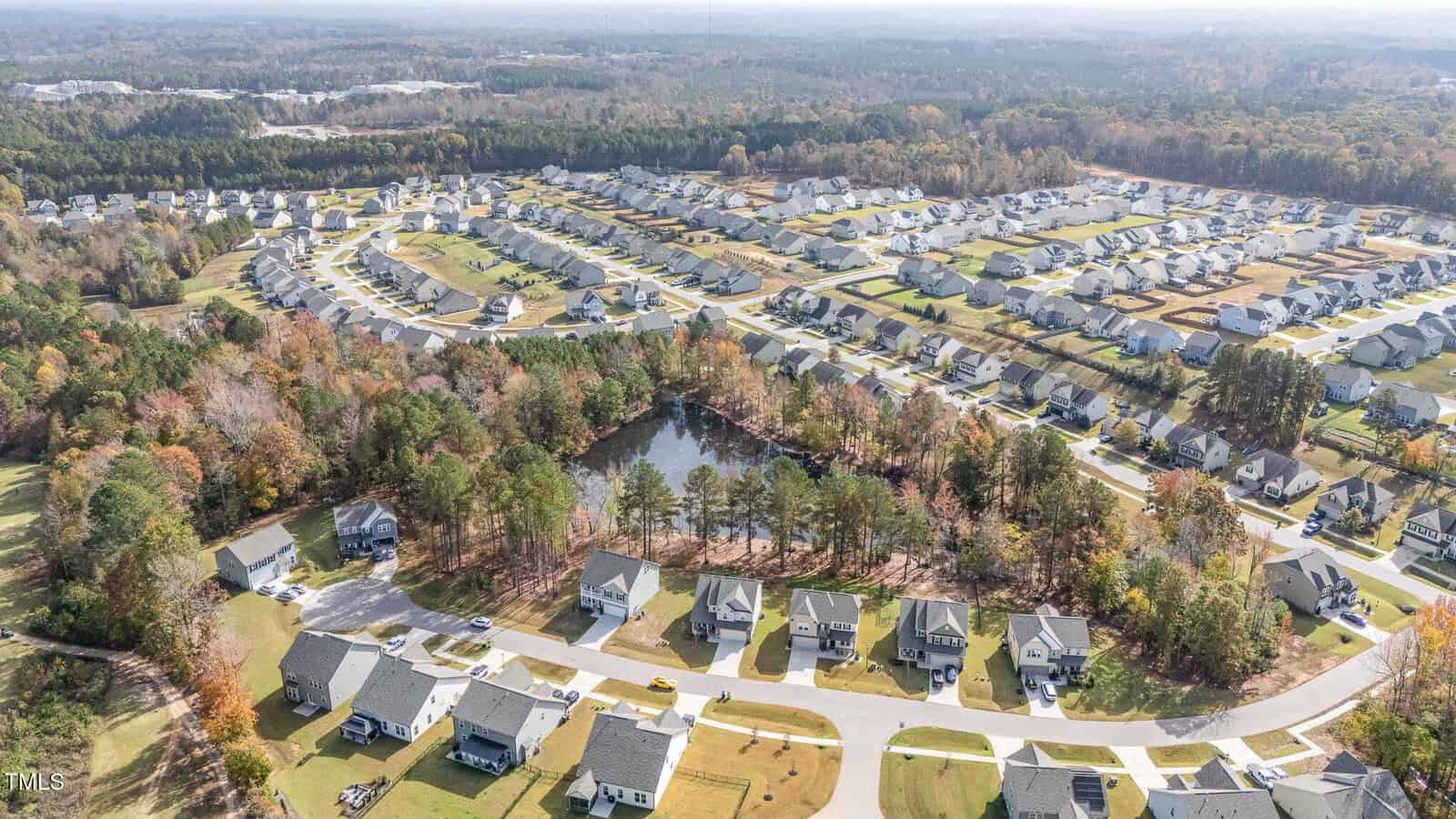 Aerial view of a suburban neighborhood with rows of houses, a small pond surrounded by trees, and expansive green lawns. The area is bordered by forested land and stretches into the distance.