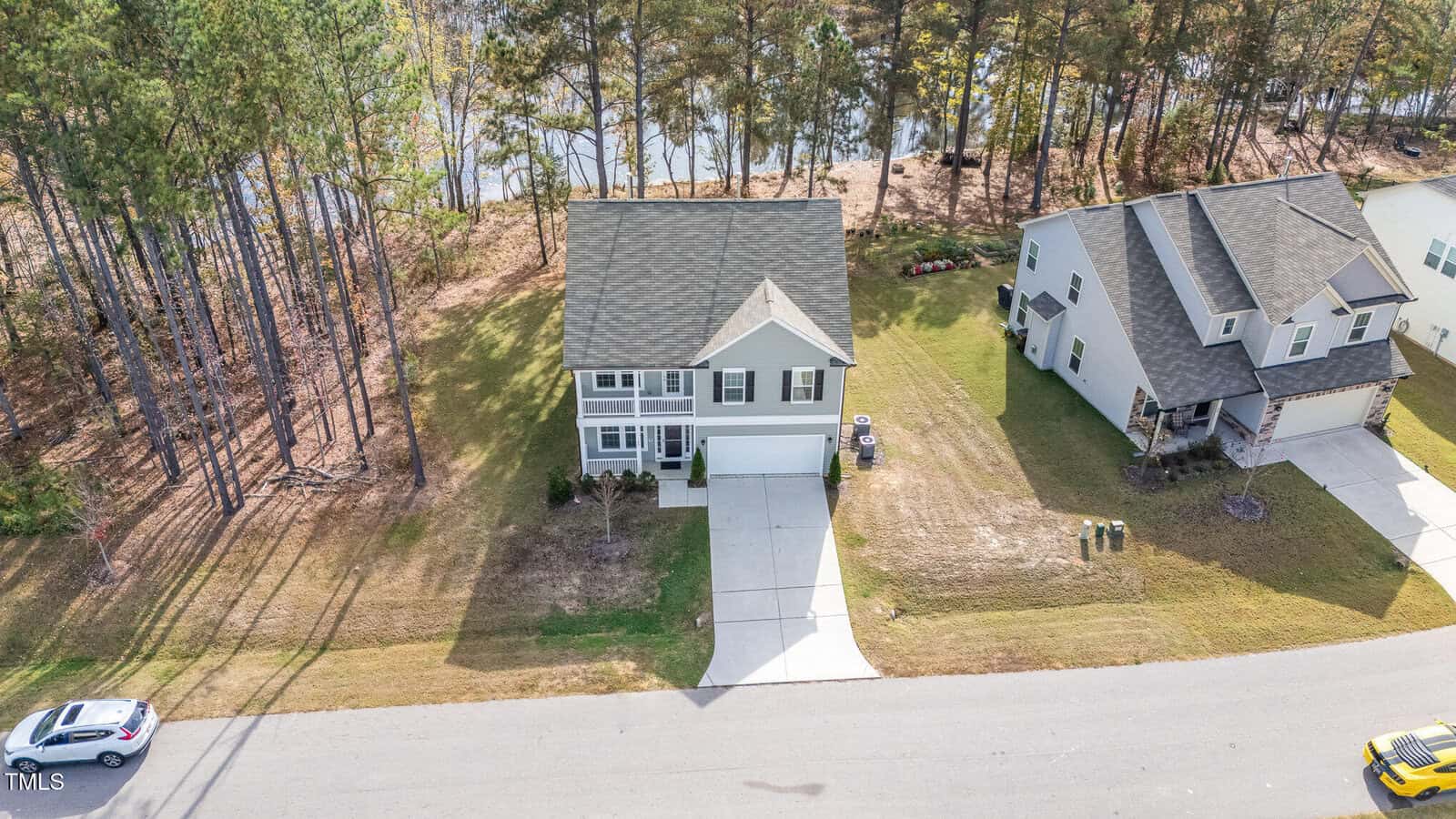 A two-story house with a gray roof and light-colored exterior sits between a wooded area and another house. It has a driveway, small front yard, and is located on a residential street. Trees and a body of water are in the background.