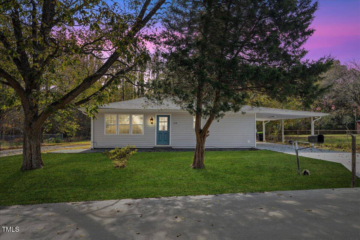 A charming single-story white house with a blue door sits under a purple and pink sunset sky at 1830 Garland Street in Henderson. It features a covered carport, surrounded by grassy lawn and trees, with a welcoming driveway leading up to the home.